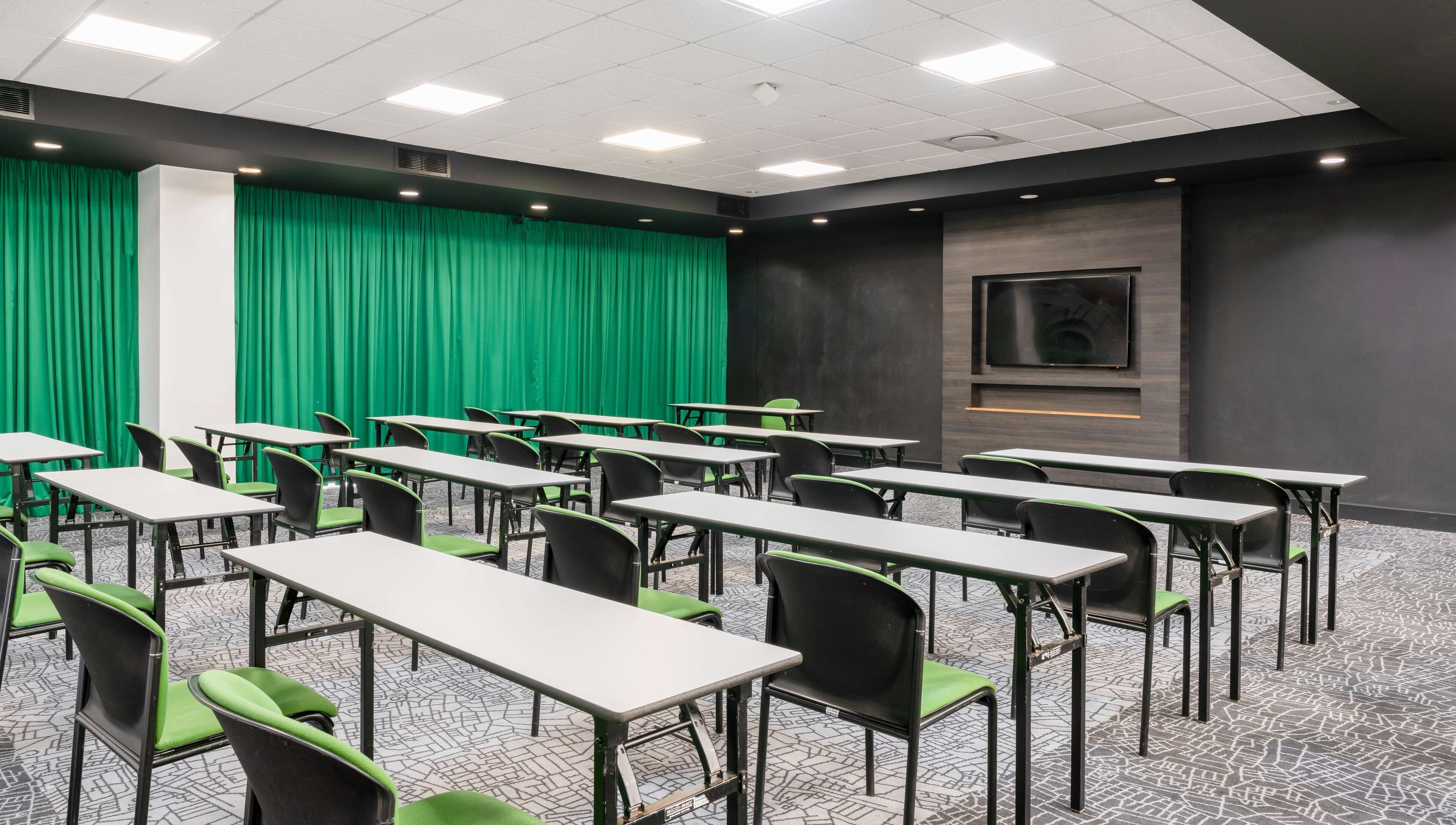 Ett konferensrum med bord och stolar, en grön bakgrund av draperad textil och en upphängd skärm på väggen.