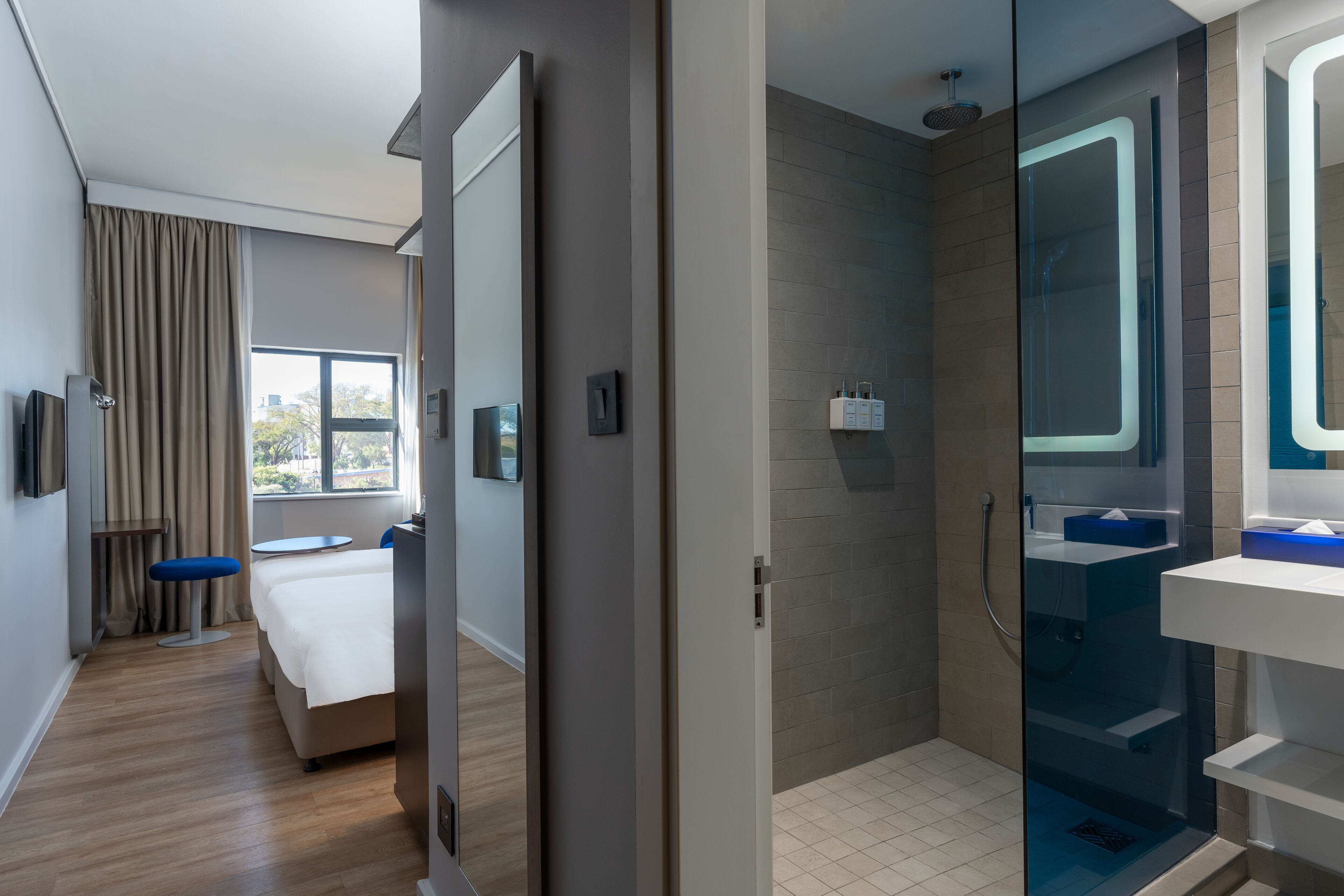 Een moderne hotelkamer met een bed, een klein tafeltje, een blauwe stoel, een groot raam met gordijnen, een douchecabine met glazen wanden en een witte wastafel.