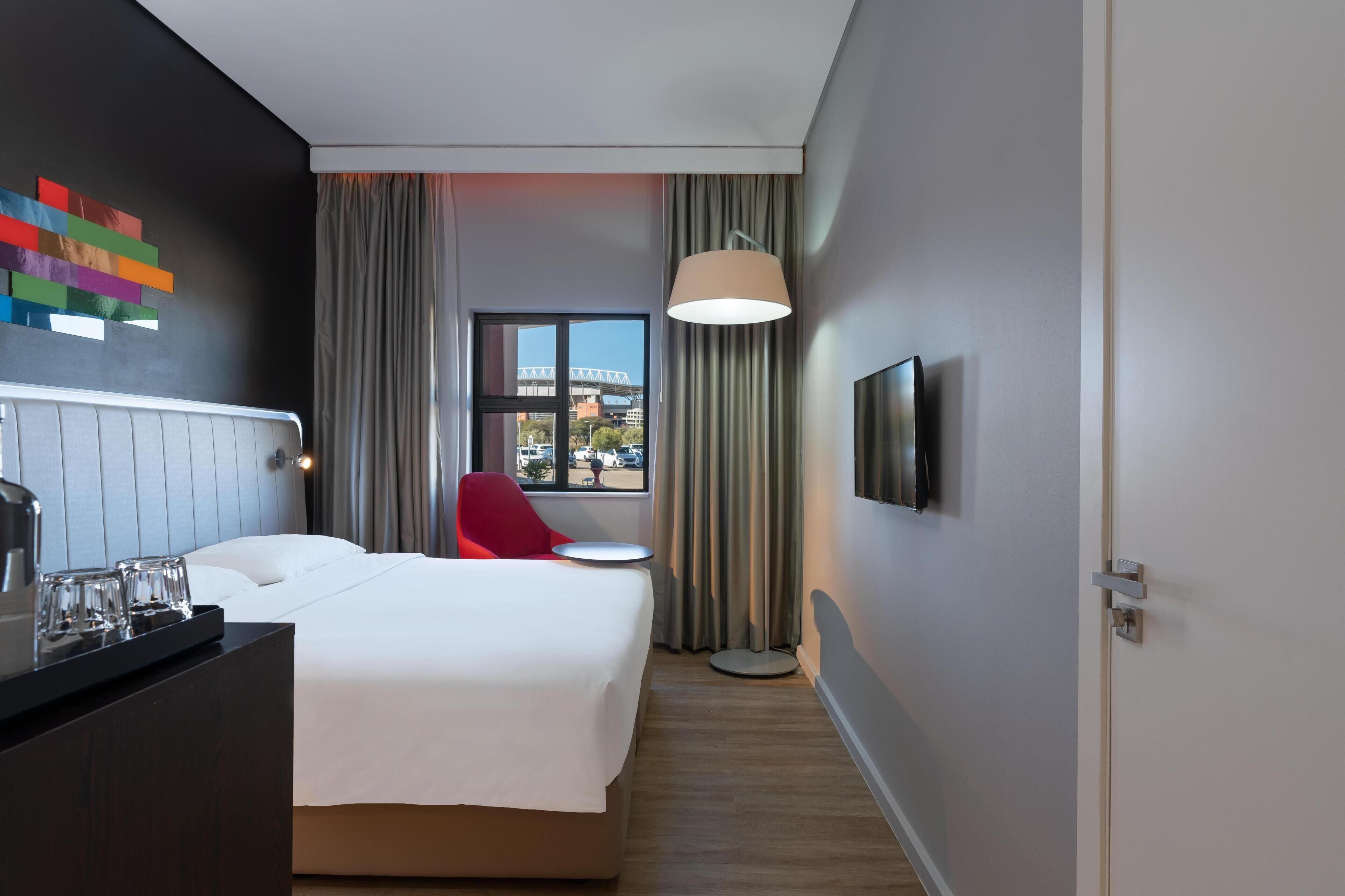 A hotel room with a bed, a red chair, a TV, and a window with a view of a bridge.