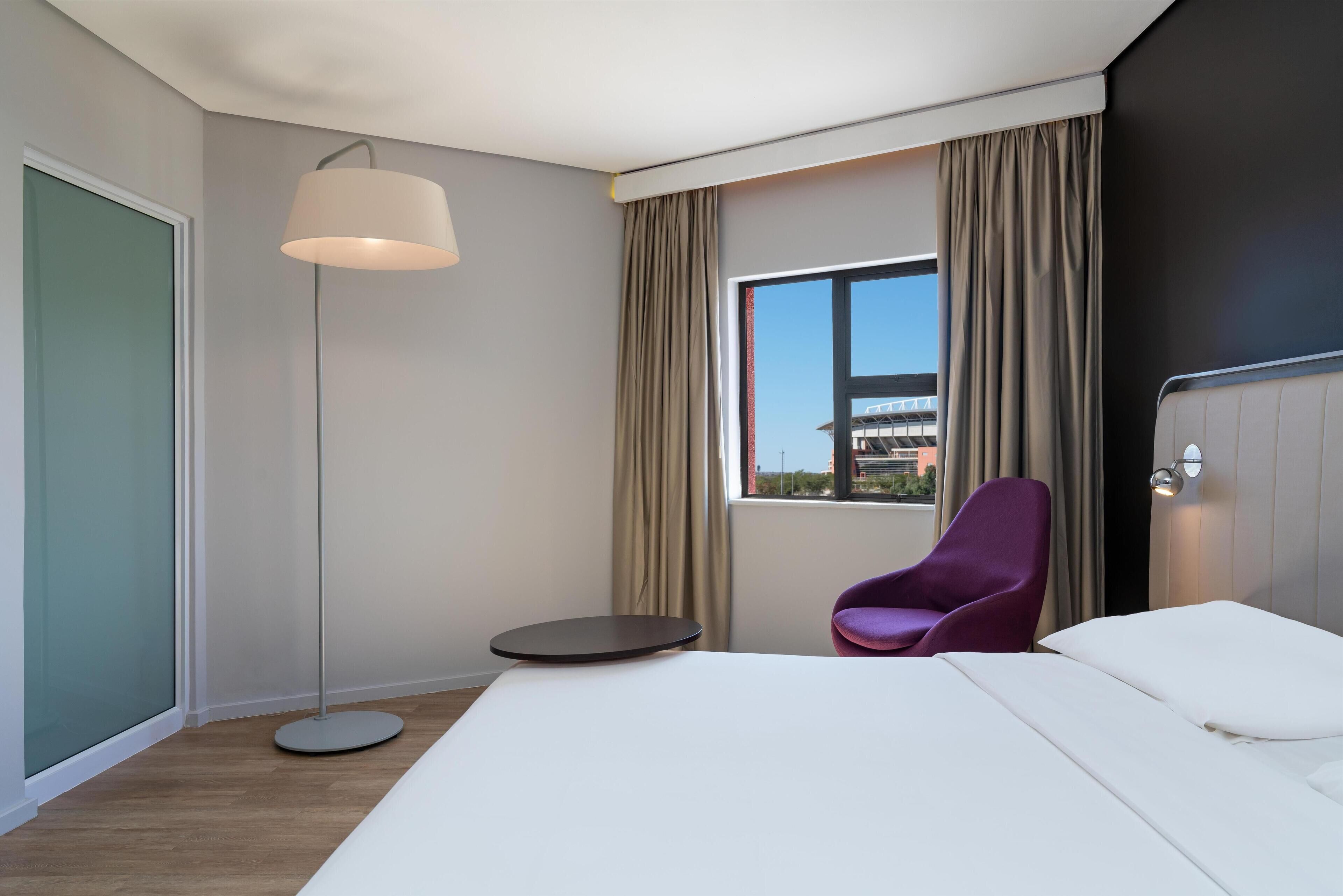 Una habitación de hotel moderna con una cama grande, una mesita redonda, una silla morada, una lámpara de pie y una ventana con cortinas.