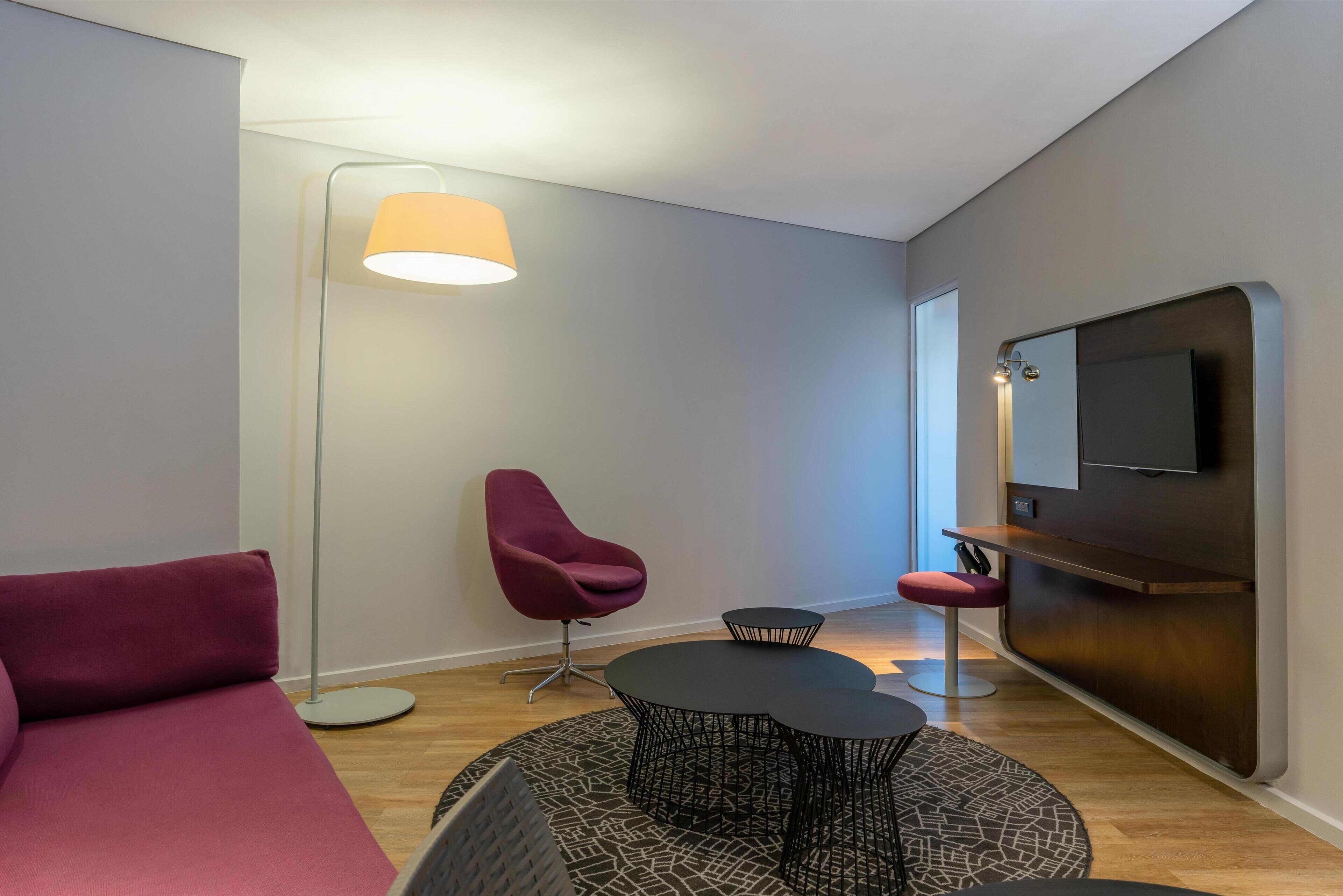 A modern living room with a sofa, a red chair, a small round table, a floor lamp, and a TV mounted on a wooden panel.