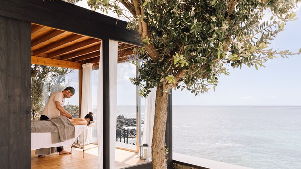 A spa room with a wooden floor, a massage table, and a view of the sea. - White Exclusive Suite & Villas (Lagoa)