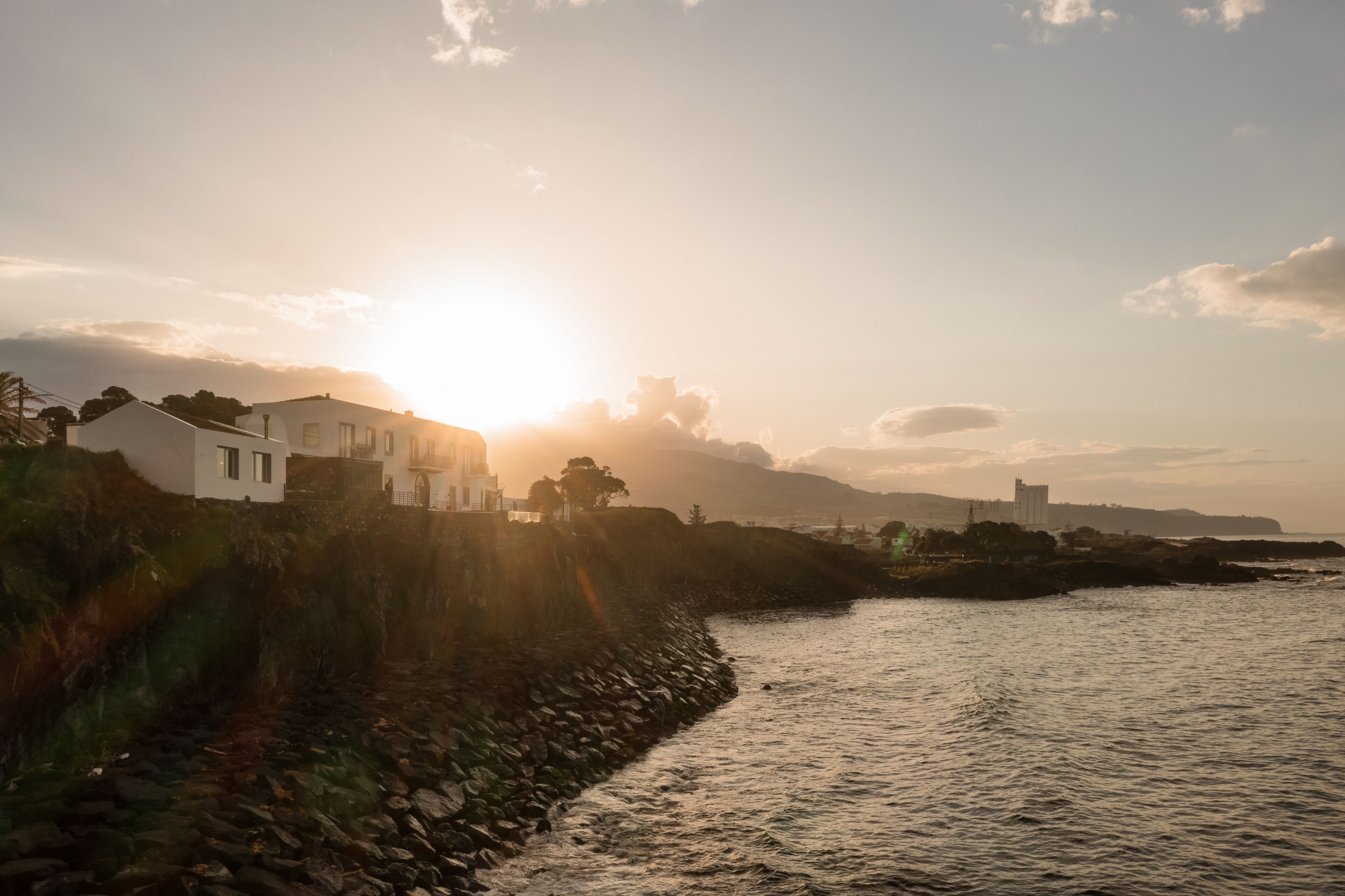 A coastal scene with a stone wall and a white building by the water during sunset.