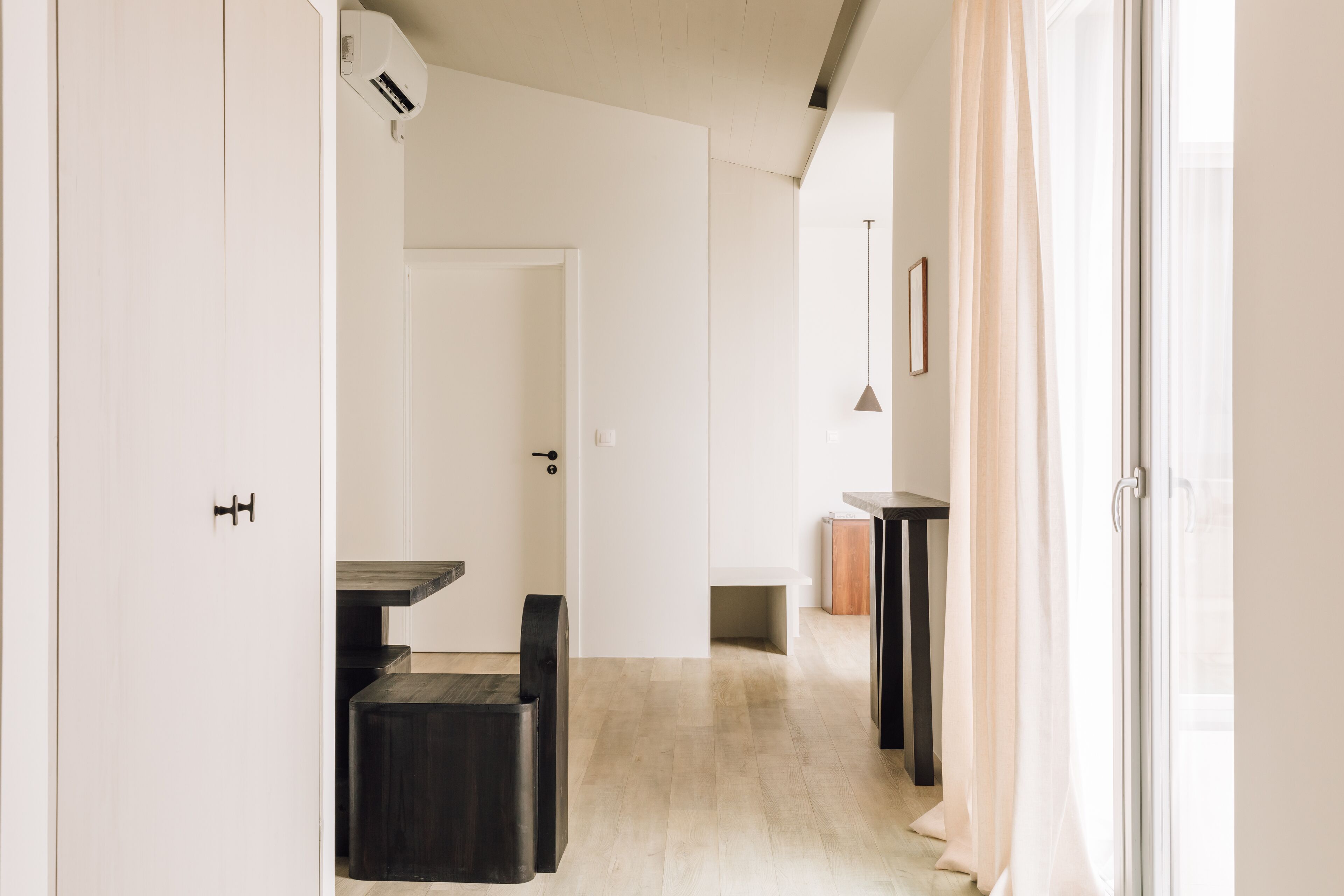 A hallway with a wooden floor, a black bench, a white door, and a small table with a light fixture above it.
