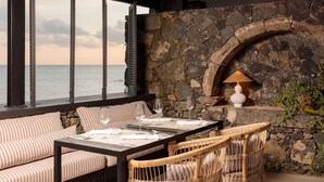 A seaside patio with a stone fireplace, wicker furniture, and a table set for dining. - White Exclusive Suite & Villas (Lagoa)