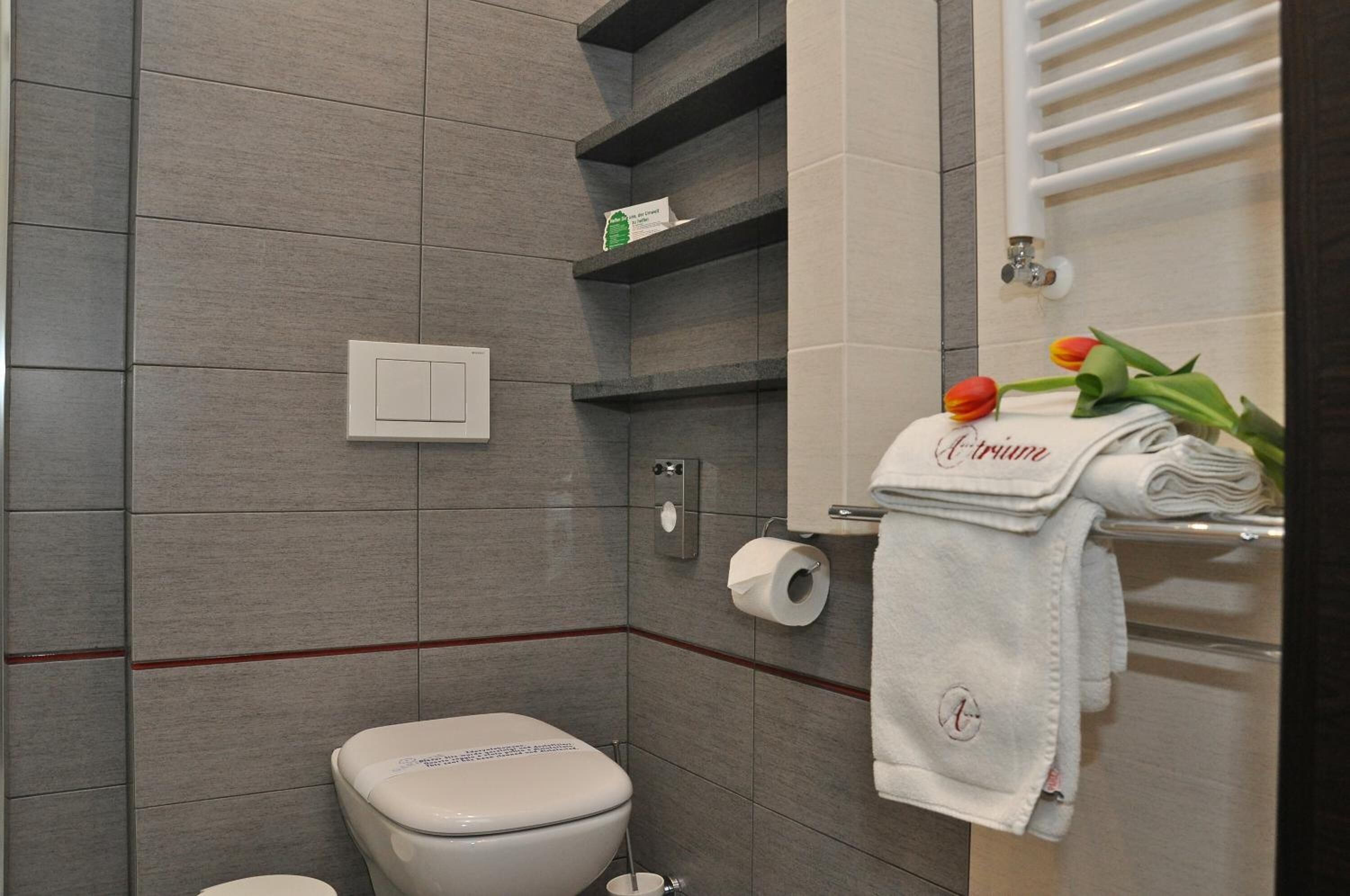 A bathroom with grey tiles, a toilet, a towel rack with a folded towel, and a toilet paper holder.
