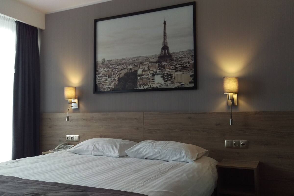 Habitación de hotel con una cama grande, dos lámparas de noche, un cuadro con la Torre Eiffel y un cabecero de madera.