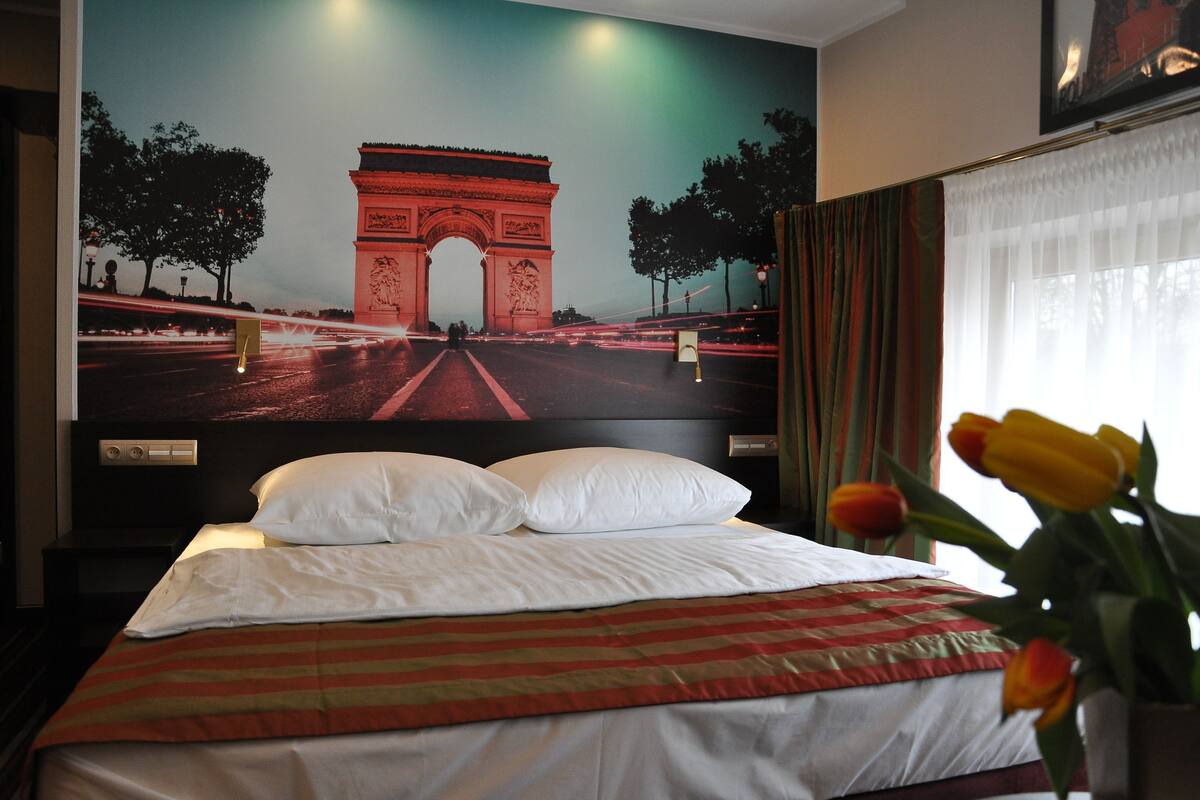 Habitación de hotel con una cama, un gran mural en la pared del Arco del Triunfo, vista nocturna y un jarrón con tulipanes.