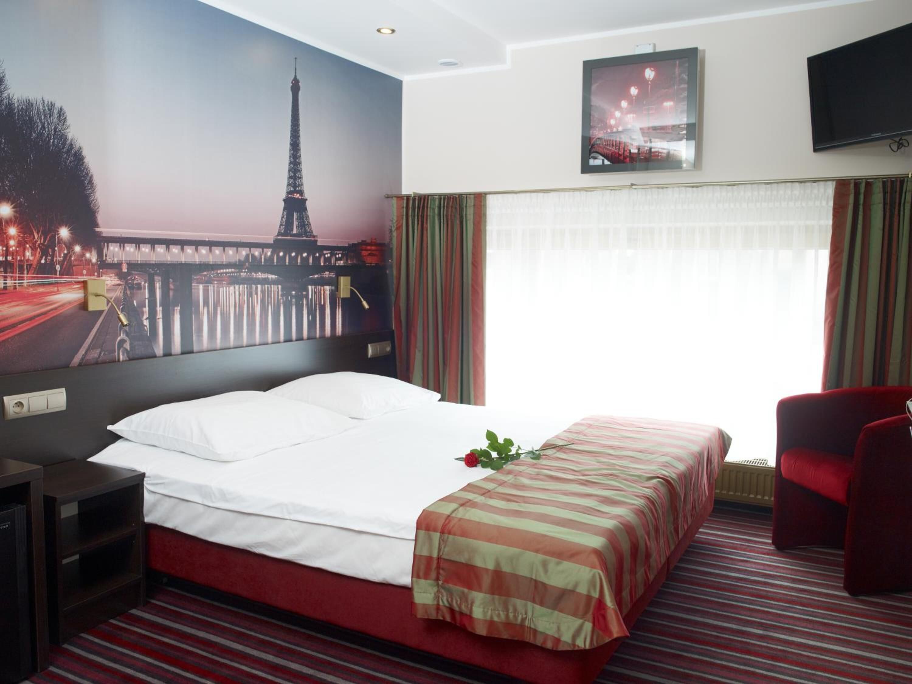 Una habitación de hotel con una cama, una silla roja, un televisor y un ventanal con cortinas.