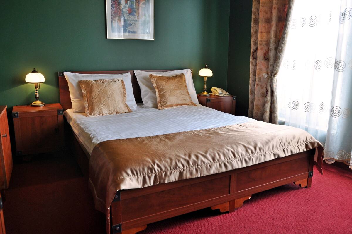 Una cama bien hecha con cubrecama marrón y dos almohadones decorativos, flanqueada por mesitas de noche de madera con lámparas, en una habitación con paredes verdes y alfombra roja.