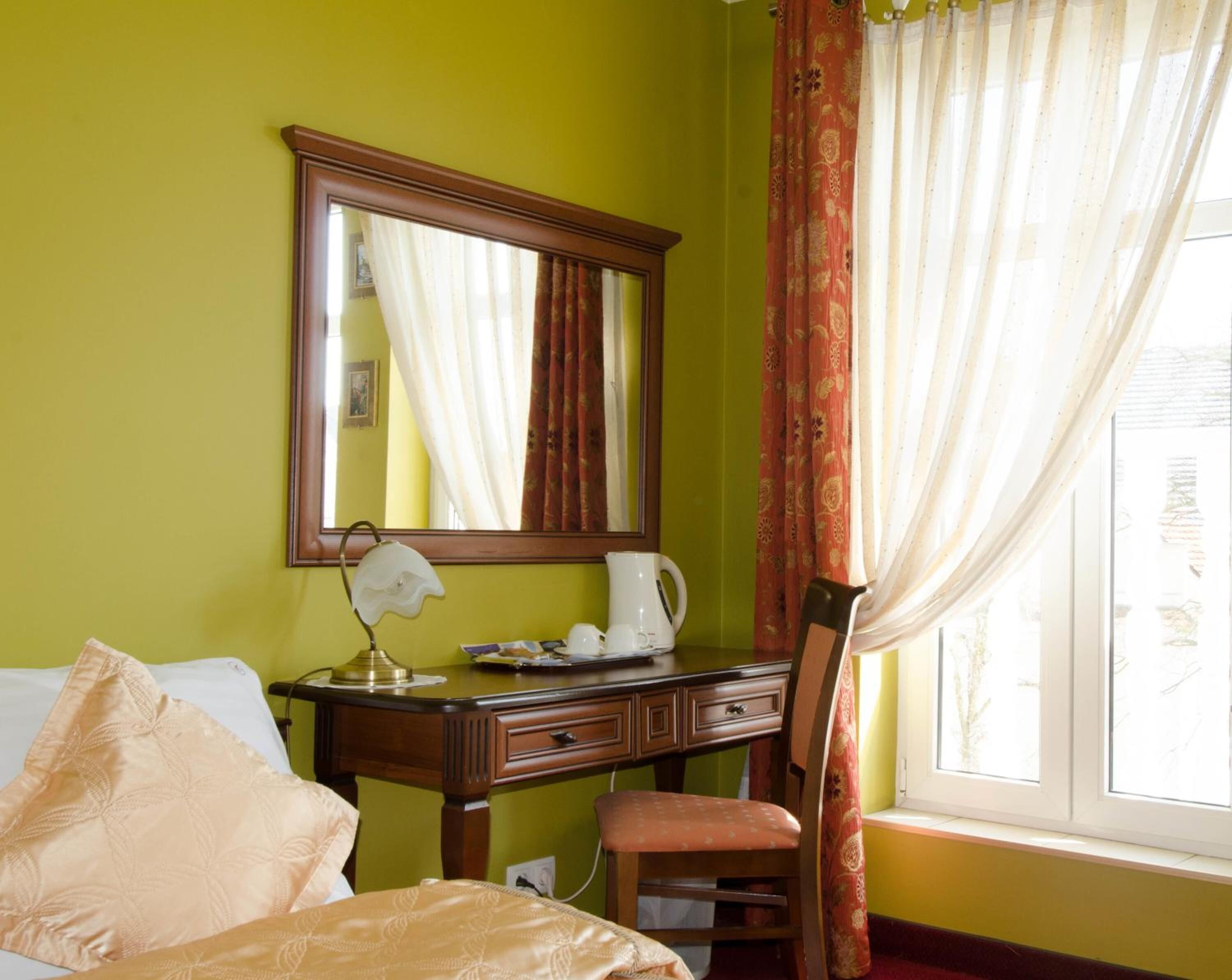 Une chambre d’hôtel avec un lit, un bureau avec une lampe, une chaise, un grand miroir et une fenêtre avec des rideaux.