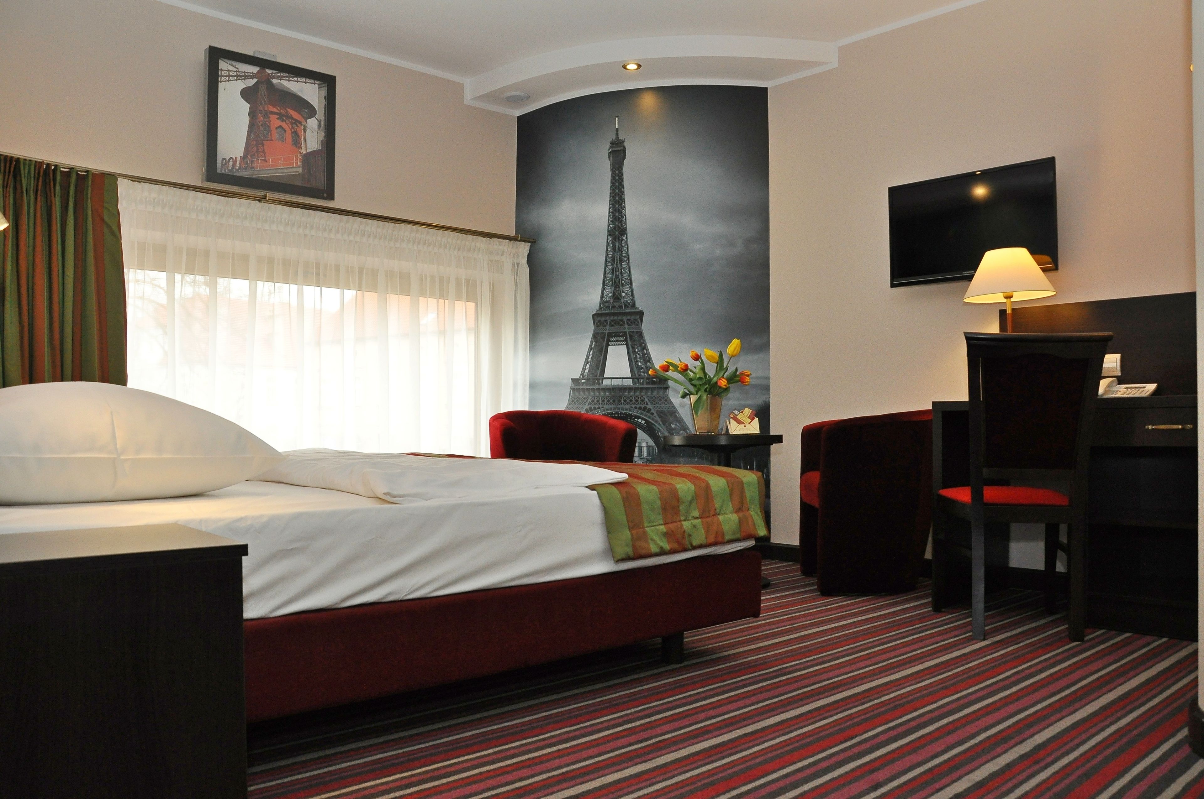 Habitación de hotel con una cama, un escritorio, una silla, un televisor y un gran mural de la Torre Eiffel en la pared.