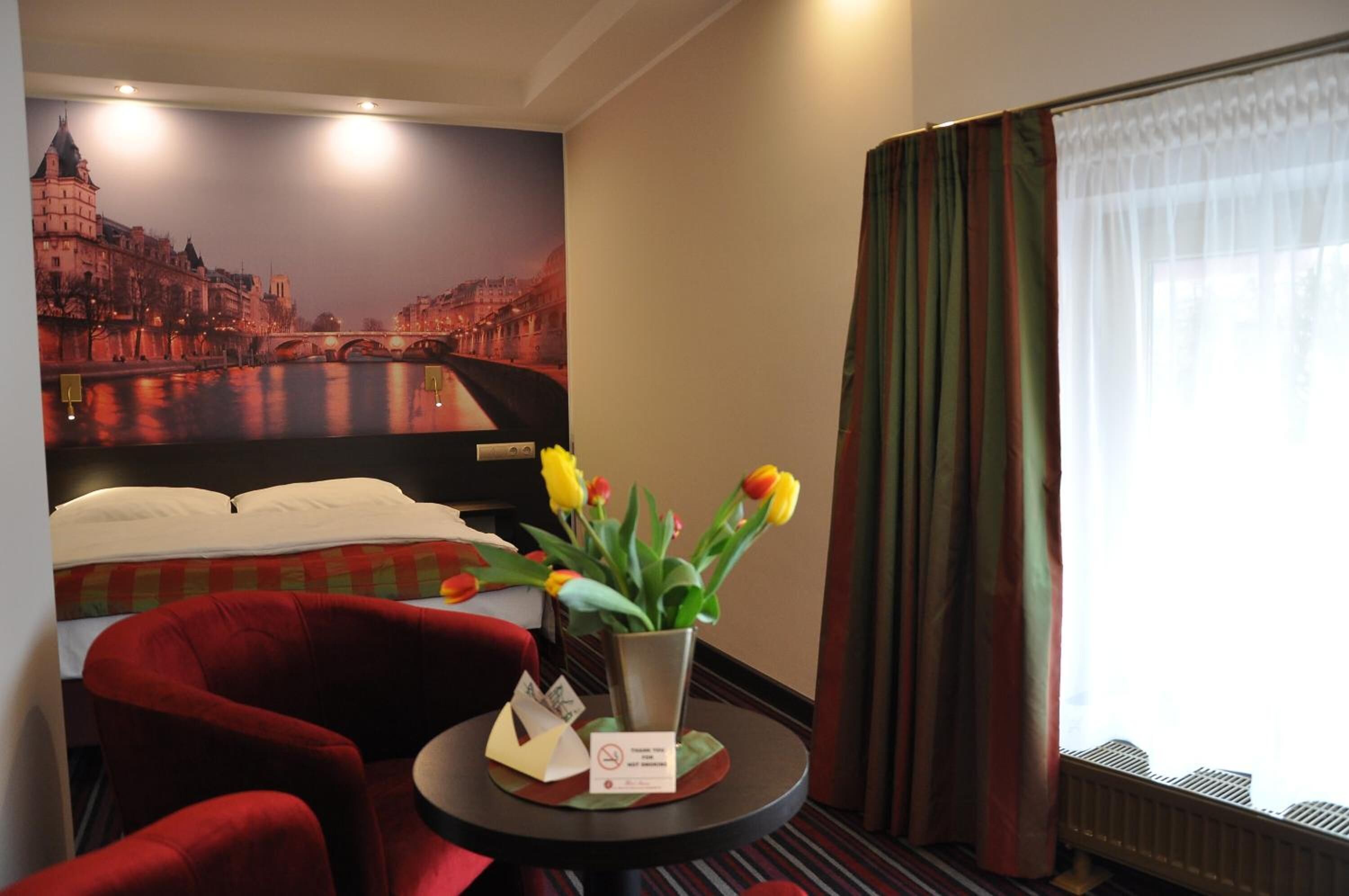 A hotel room with a bed, a red armchair, a small table with a vase of tulips, a poster of a cityscape, and a window with curtains.