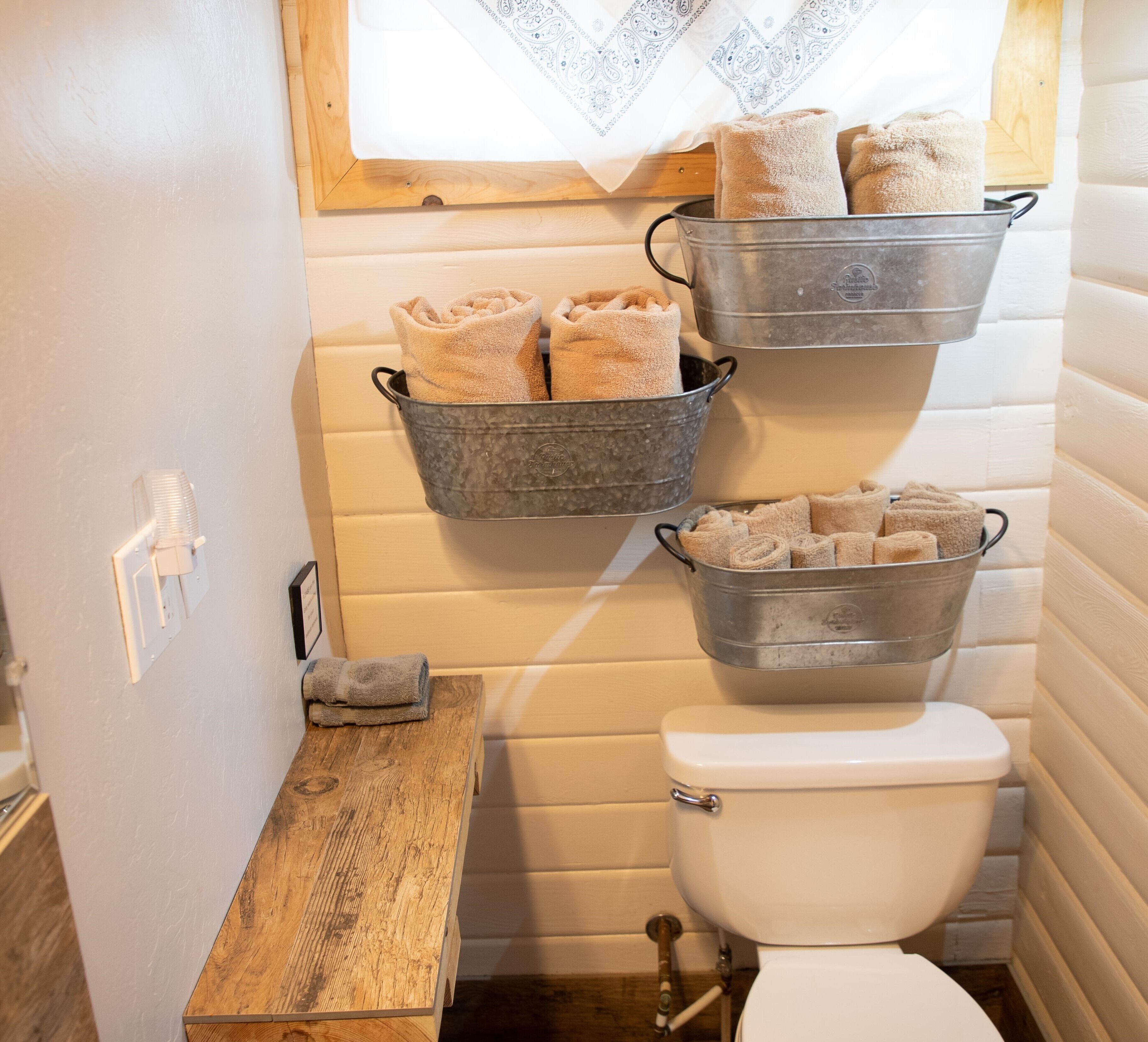 Un baño pequeño con un inodoro, un banco de madera y cubos metálicos con toallas.