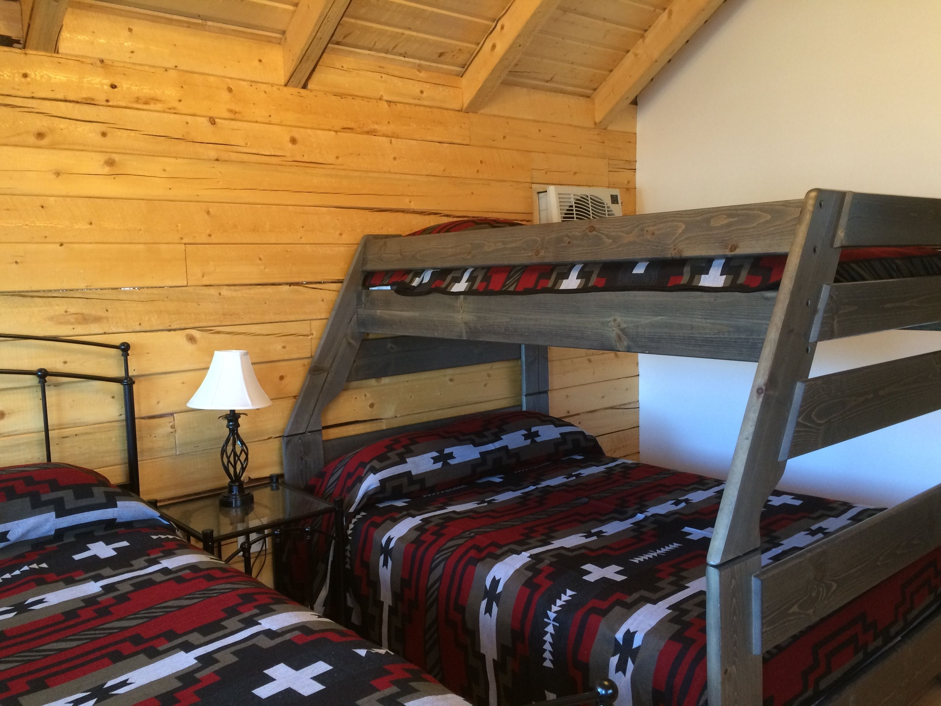 Ein Schlafraum mit Etagenbetten, Holzboden und -wänden, einem Nachttisch mit Lampe sowie einem Bett mit Musteroptik.