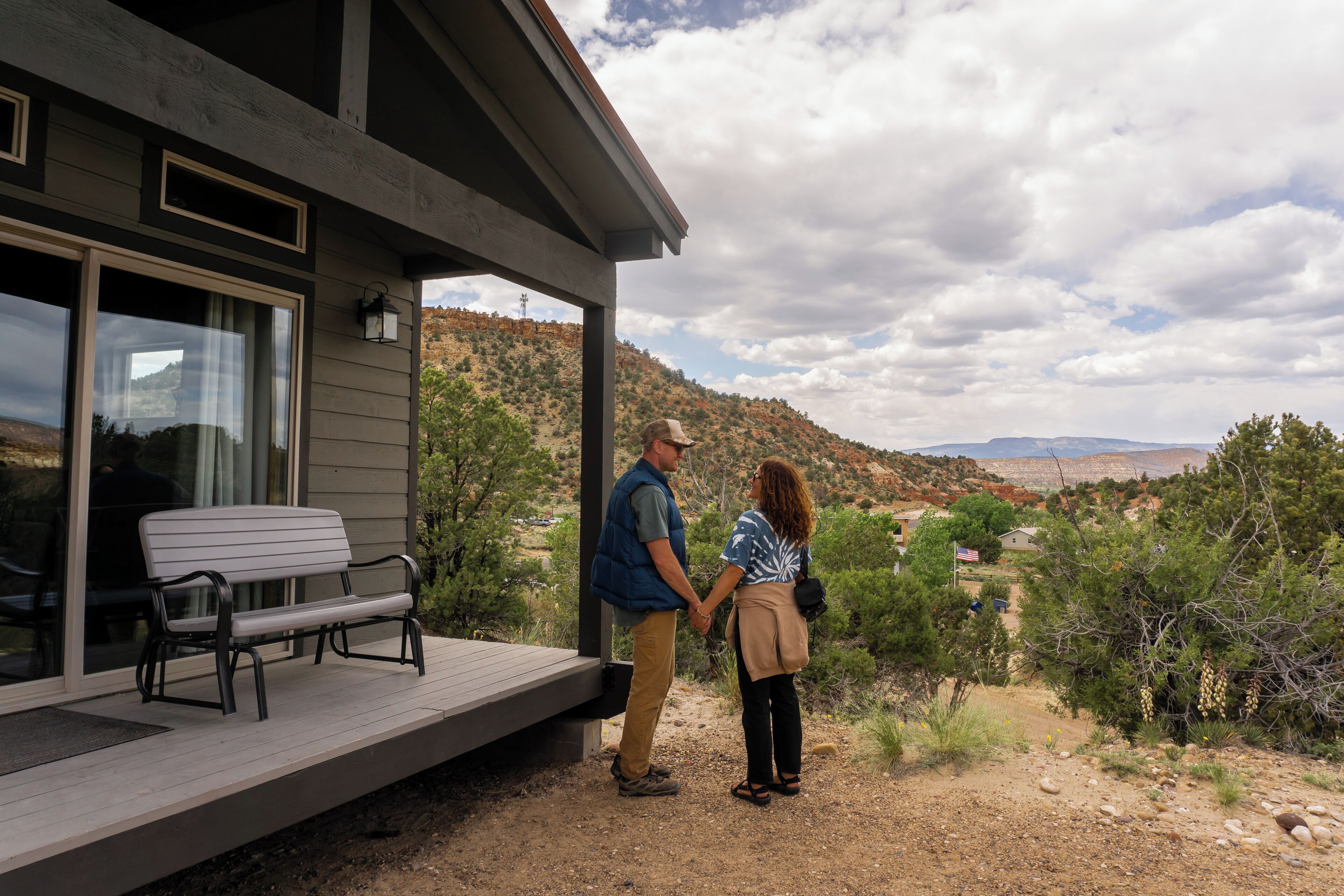 A couple holding hands on a wooden deck with a bench, overlooking a hilly landscape with sparse vegetation and a clear sky.