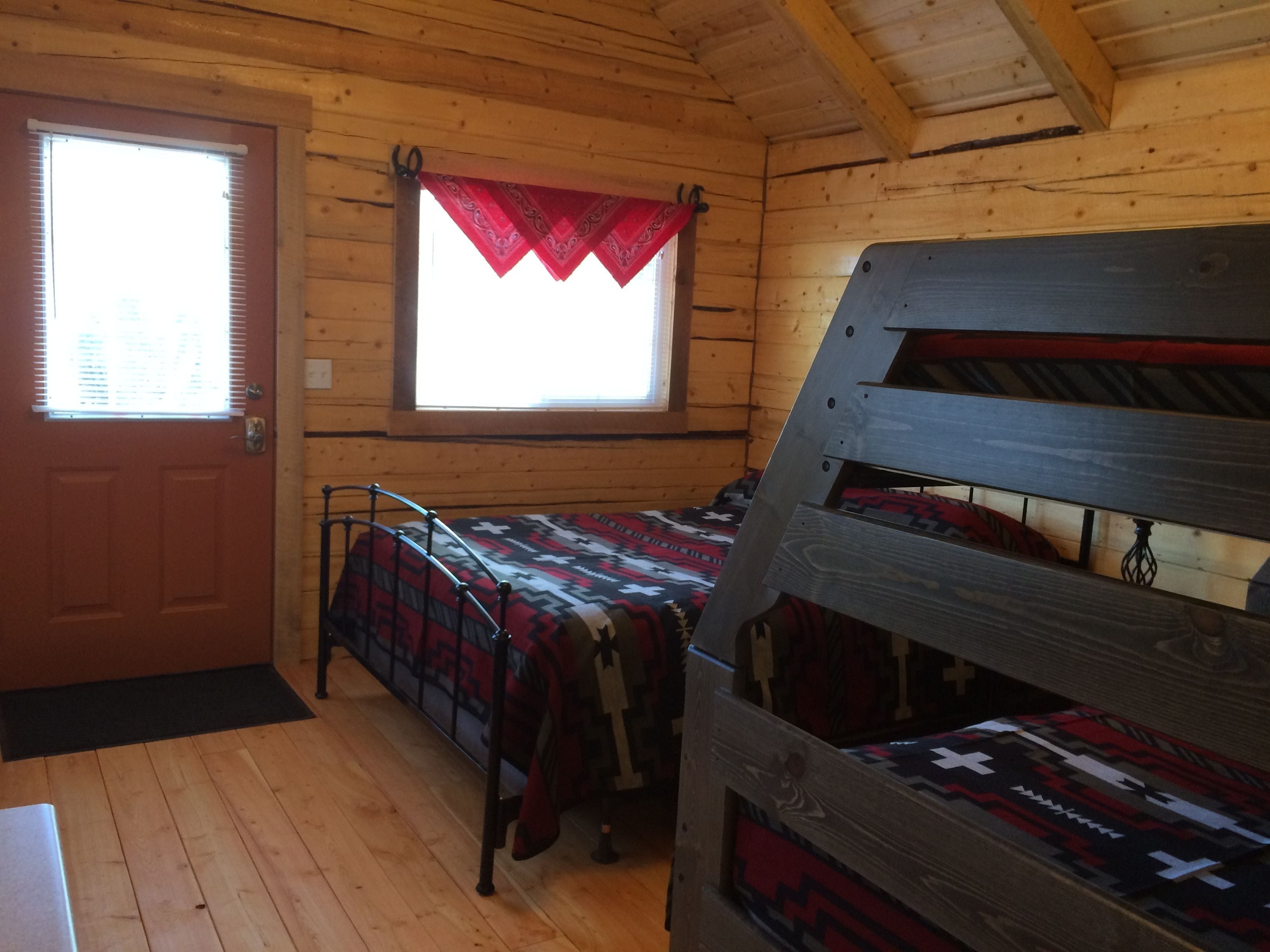 Mökkityyppinen huone, jossa on puinen sisustus, kerrossänky, punaisella reunuksella varustettu ikkuna ja sälekaihtimilla varustettu ovi.