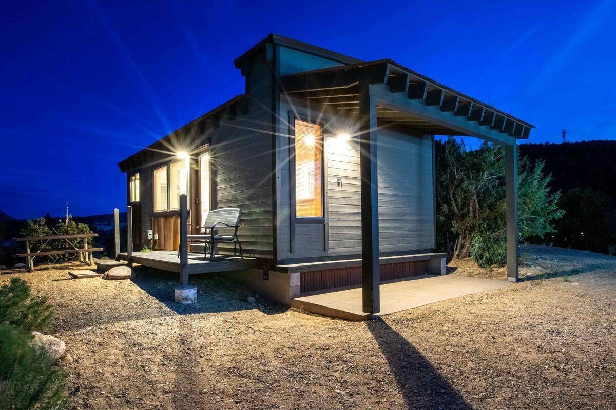 A modern, two-story cabin with a covered porch and a small deck, surrounded by a gravel driveway and sparse vegetation.