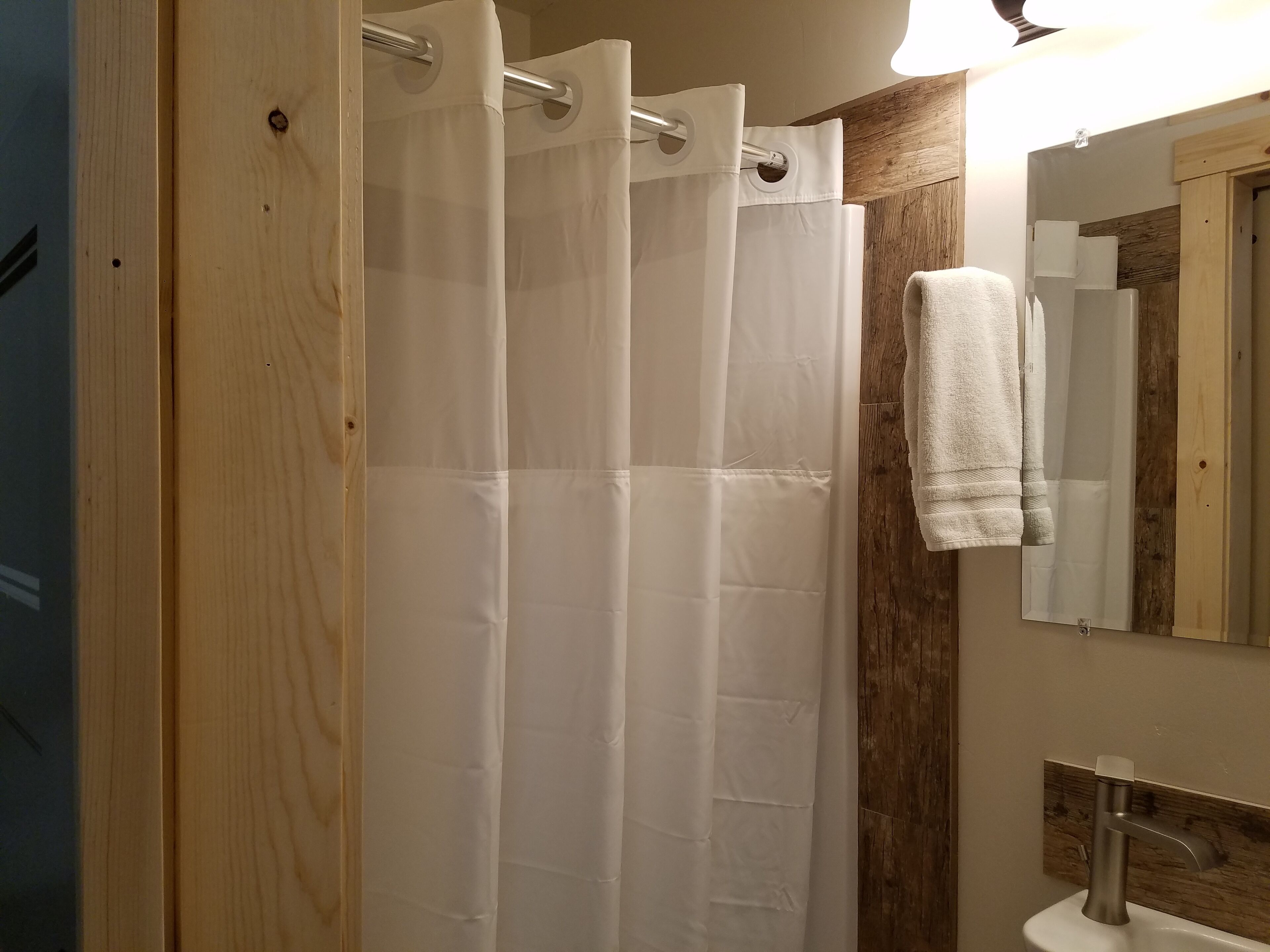 Une salle de bain avec une porte en bois, un rideau de douche blanc, une serviette accrochée au mur et un miroir au-dessus d’un lavabo.
