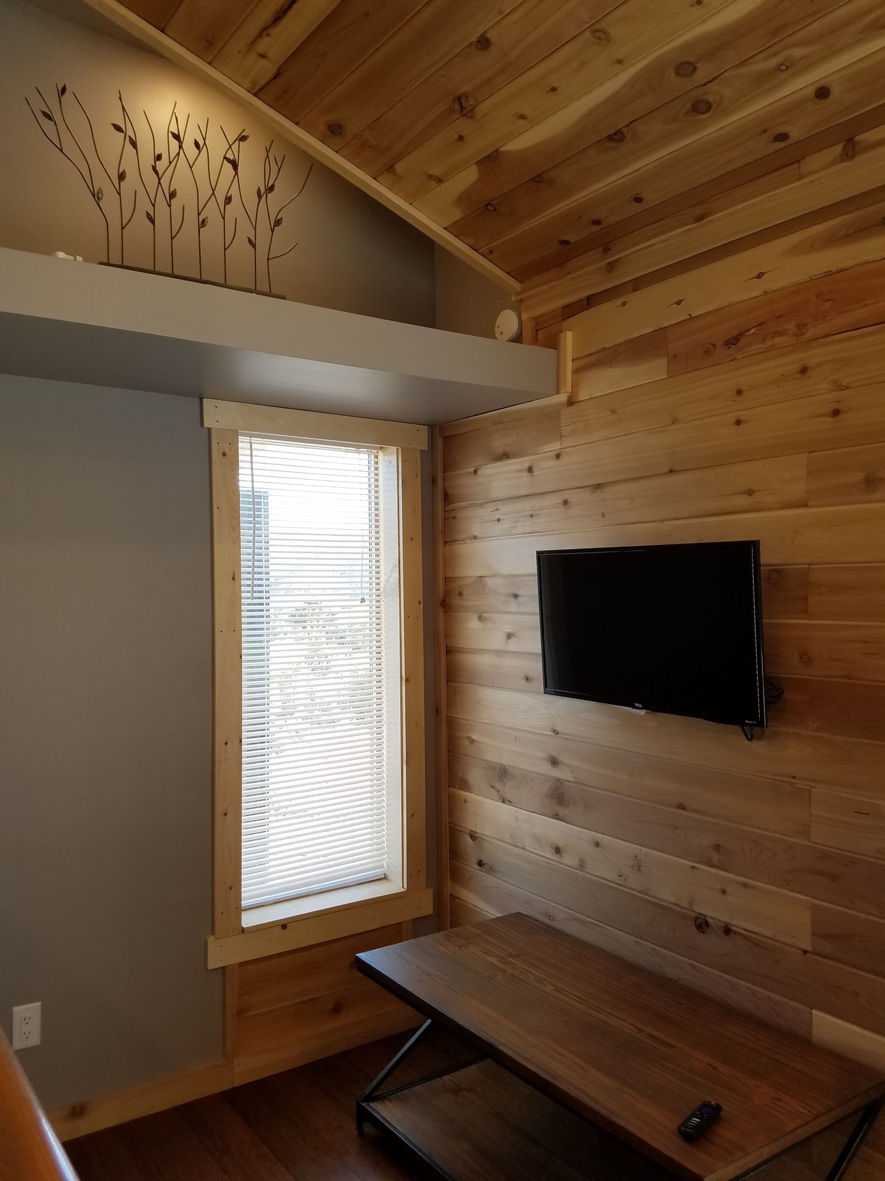 A room with wooden panel walls, a window with blinds, a flat-screen TV mounted on the wall, and a foldable bench.