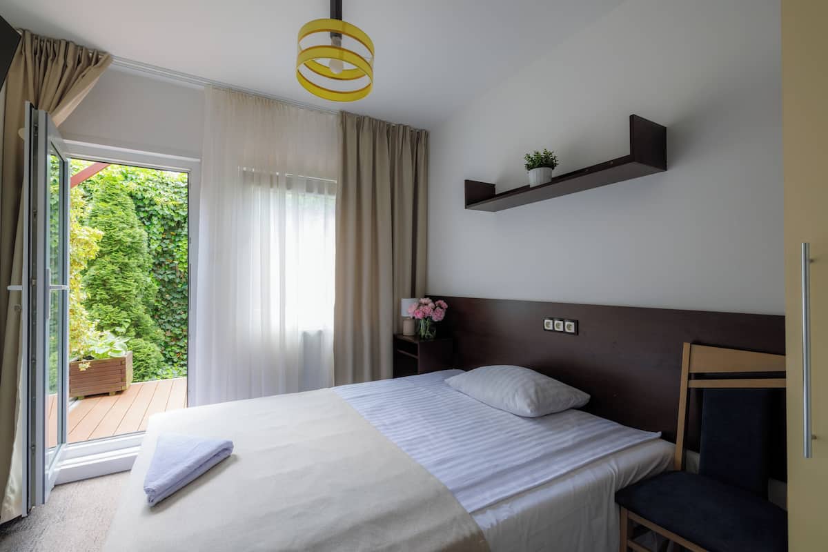 Habitación de hotel con una cama, una silla, una mesita de noche con lámpara, un estante con una planta y vistas al exterior con vegetación.