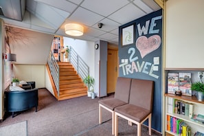 A modern lobby with a staircase, a blackboard with 'WE LOVE TRAVEL' message, a brown chair, and a bookshelf with books. - Smart2Stay Magnolia (Warsaw)