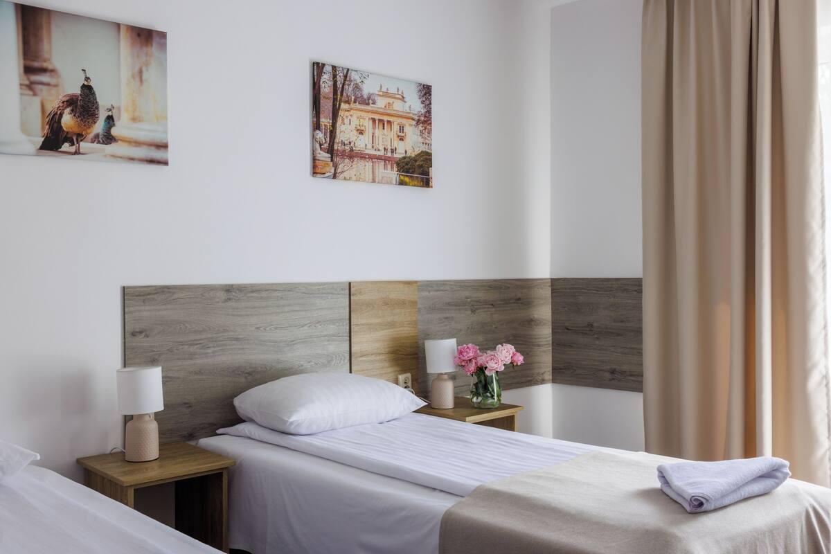 A hotel room with a wooden headboard, a bed with white linens, a nightstand with a lamp and flowers, and two framed pictures on the wall.