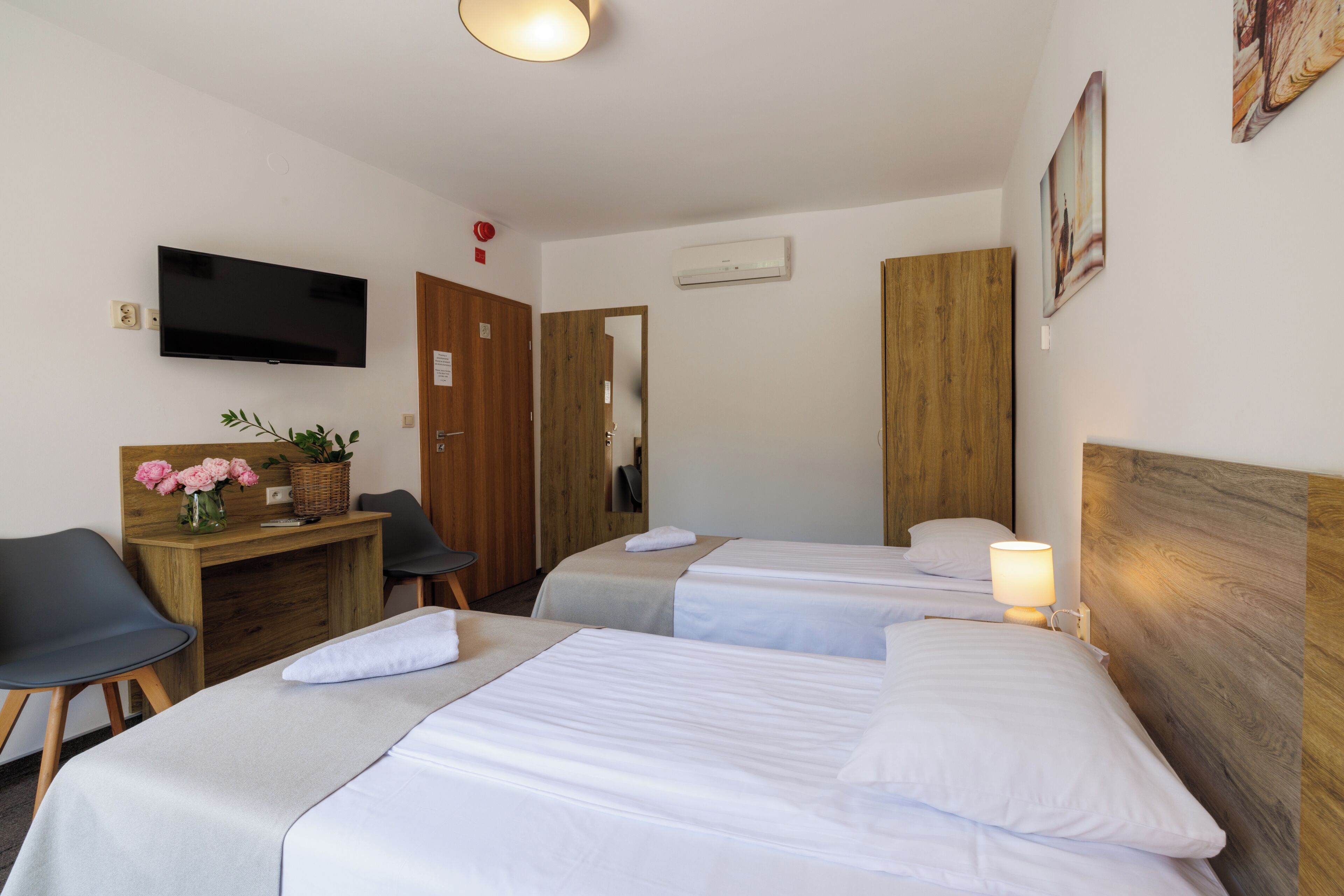 A hotel room with two beds, a desk with a chair, a television, and a wall-mounted air conditioner.