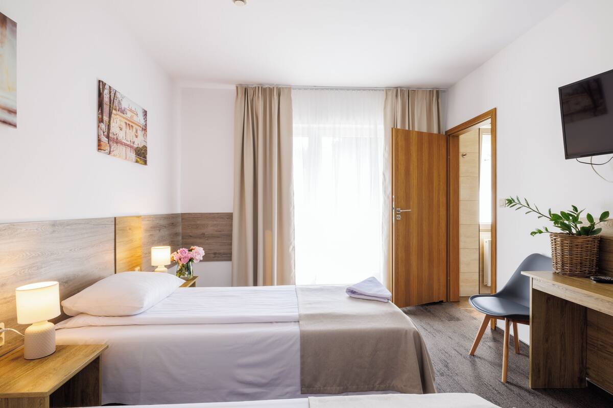 A hotel room with a bed, bedside tables, a desk, a chair, a television, and a window with curtains.