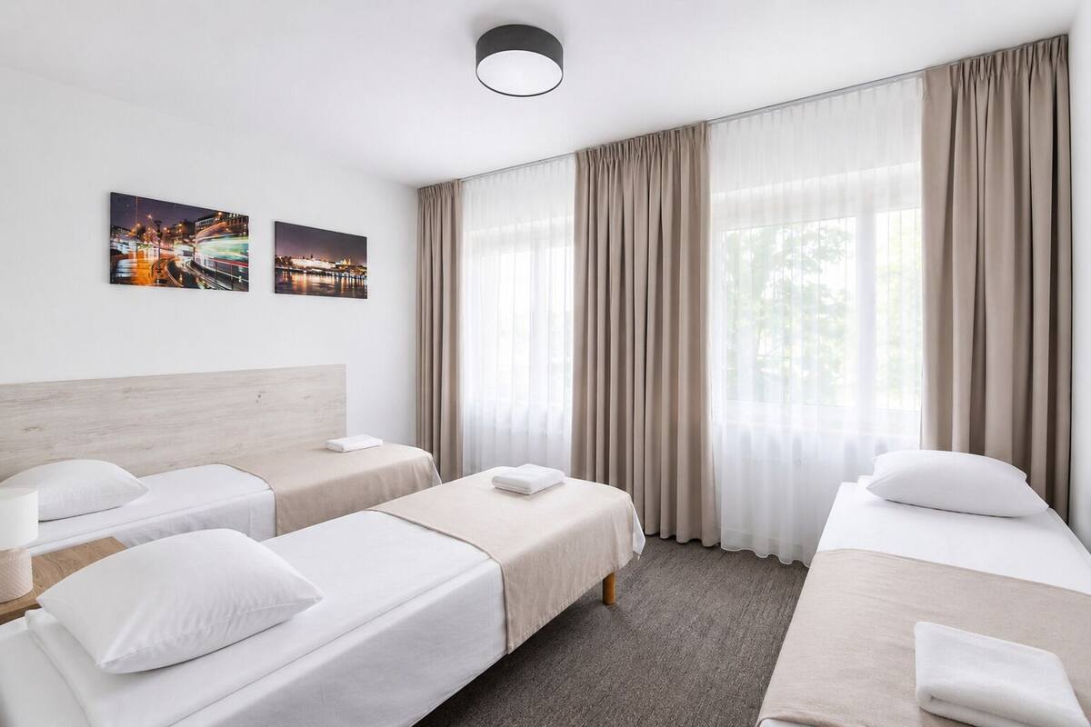 A hotel room with two single beds, a wooden headboard, a large window with curtains, and two wall-mounted pictures.