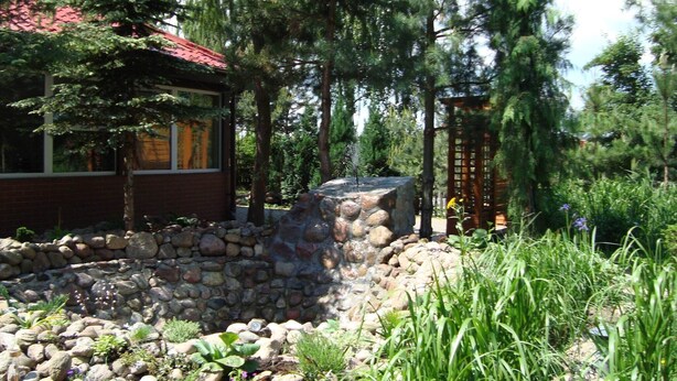 A rustic cabin with a stone foundation, surrounded by dense trees and a small waterfall feature. - Holiday Home Zgorzale by Lake Radunskie (Zgorzałe)