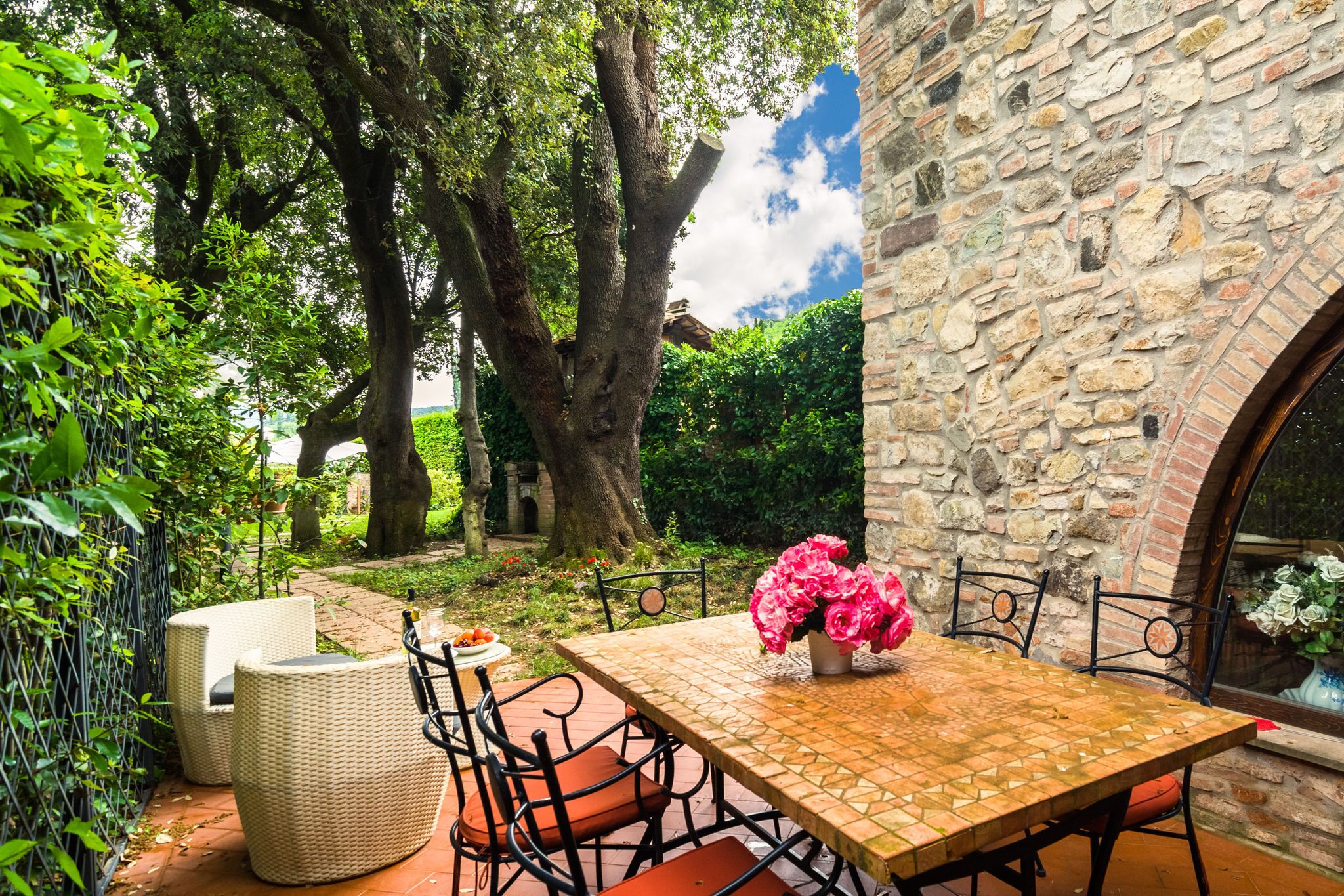 Eine Terrasse mit Steinmauer, Korbmöbeln und einem Tisch mit Blumen.