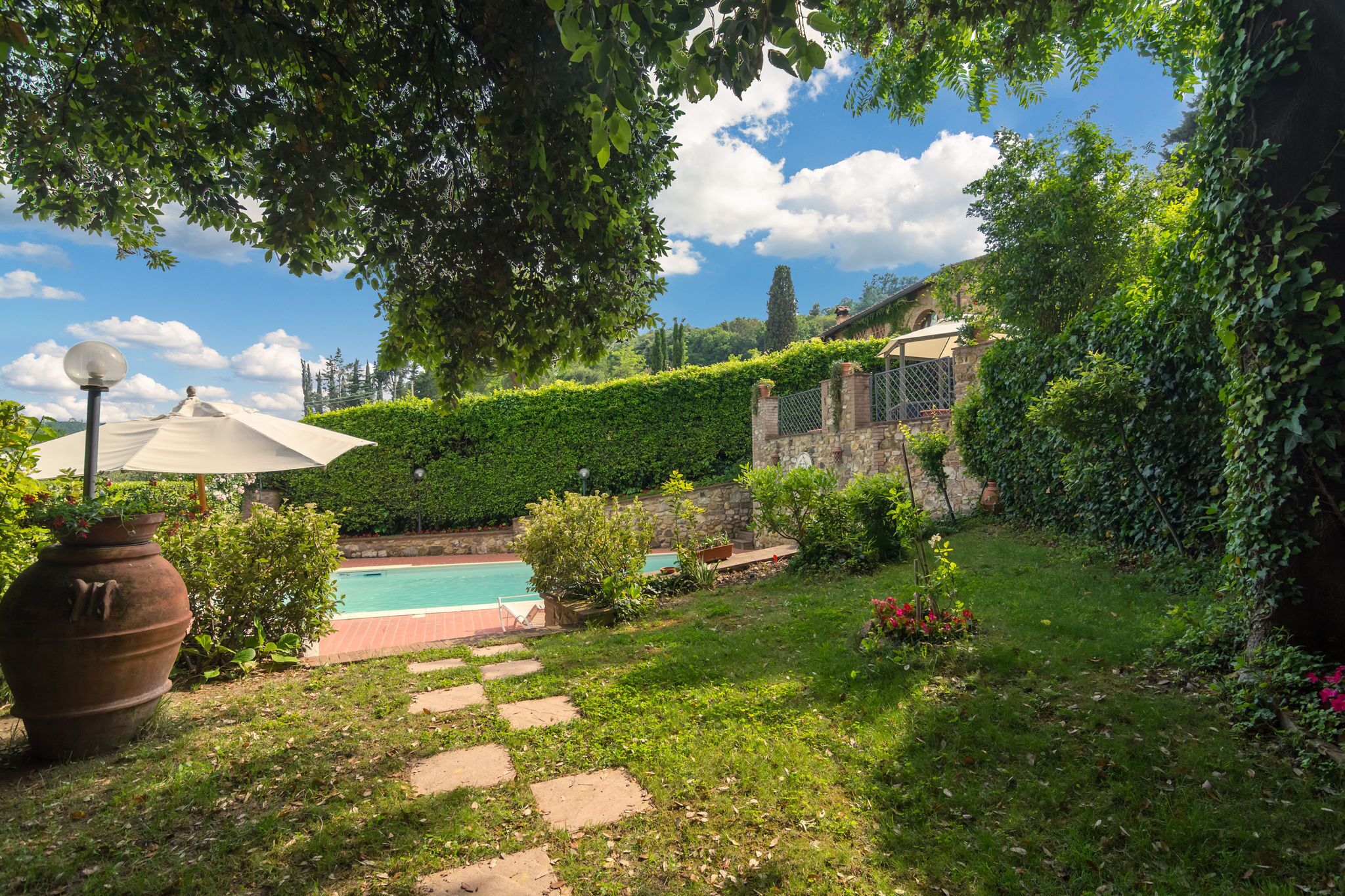 A garden with a swimming pool, stone steps, and a pergola with an umbrella.