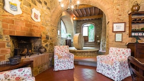 Un interior rústico de piedra con chimenea, dos sillones con motivos florales y una escalera de madera que conduce a un espacio al aire libre.