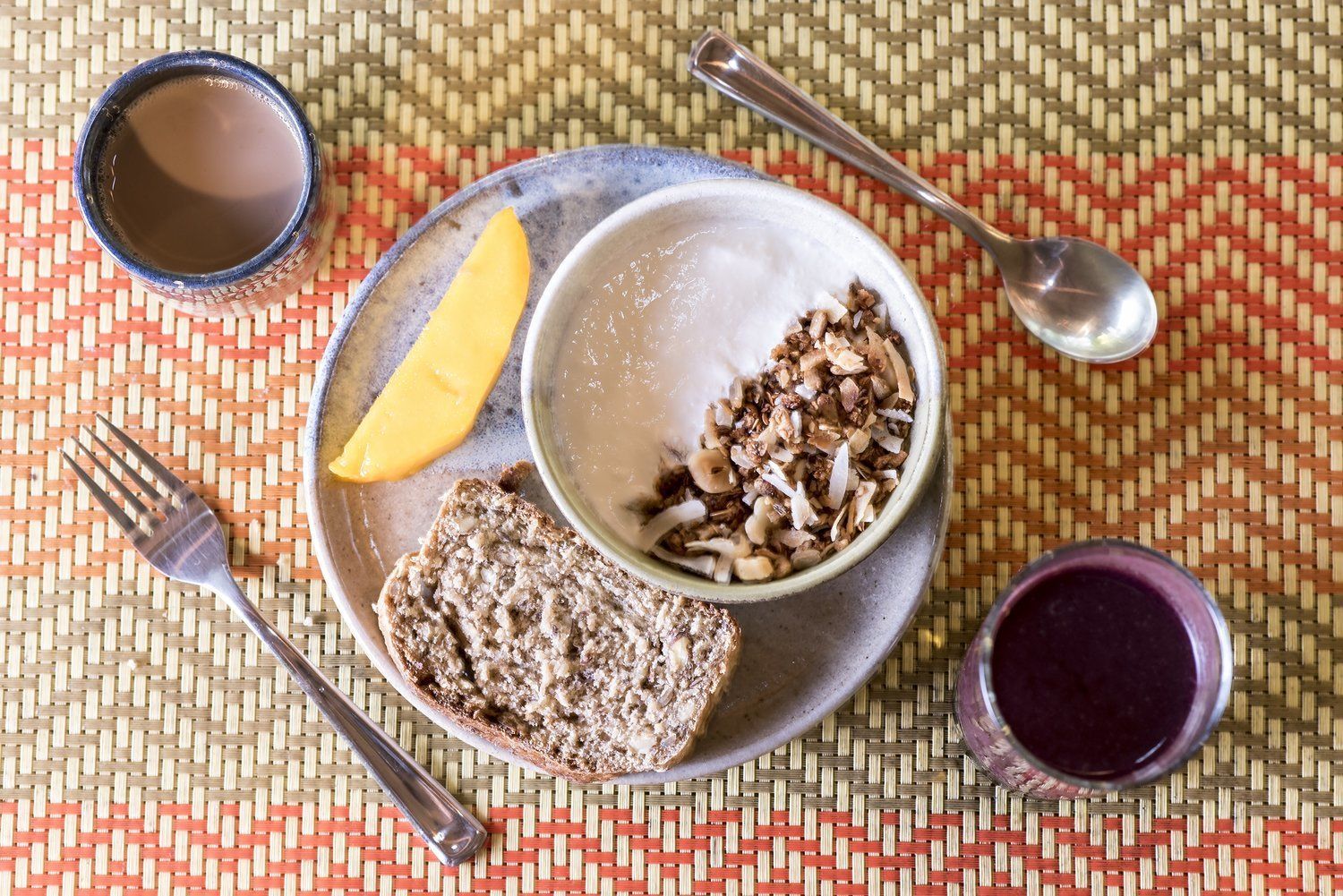 A meal with bread, granola, a slice of mango, and a cup of tea on a woven placemat.