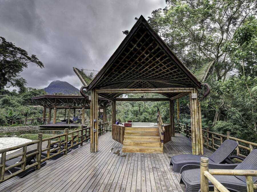 Una terraza de madera con un jacuzzi y sillones de descanso, rodeada de abundante vegetación y una montaña al fondo.