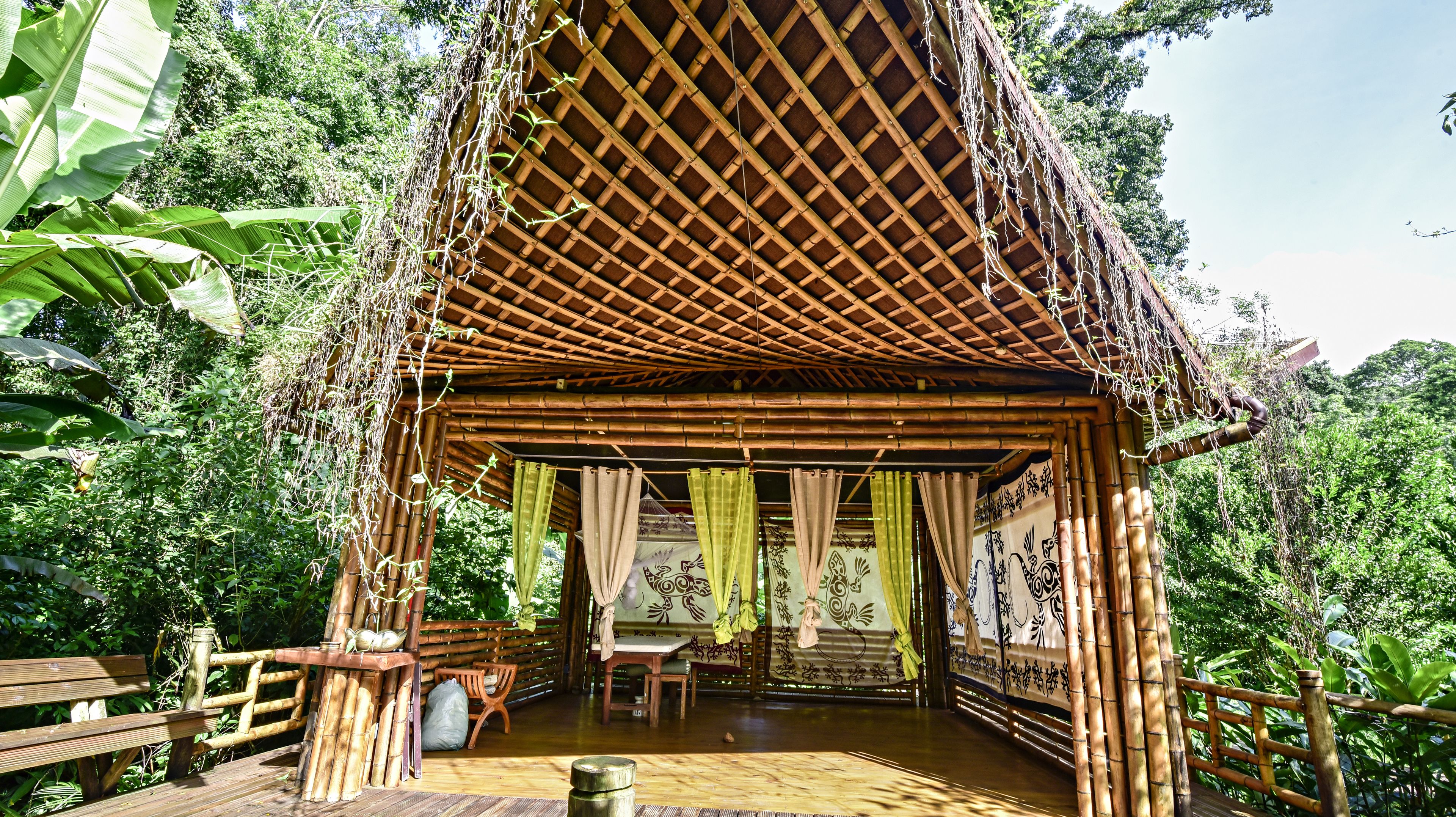 Una estructura de bambú con techo de paja, un banco de madera y una mesa con sillas en su interior.