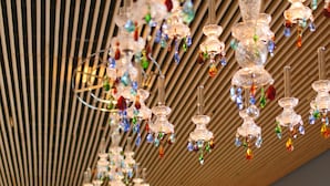 Een modern interieur met een houten spiltrap plafond en hangende kroonluchters met kleurrijke glaselementen. Er zijn zithoeken met oranje en gele kussens, en er is iemand zichtbaar op de achtergrond.