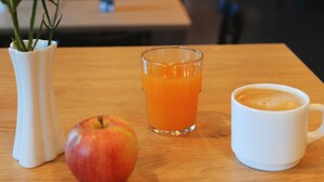 Een eettafel met een witte vaas vol bloemen, een appel, een glas jus d'orange en een witte mok.