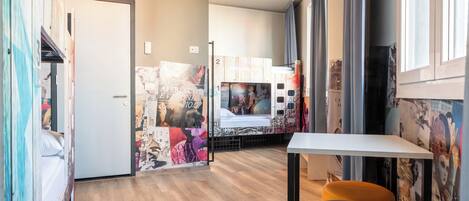 Ein Hotelzimmer mit Holzboden, einer weißen Tür, einem Bett, einem kleinen Tisch und einem Fenster mit Vorhängen.