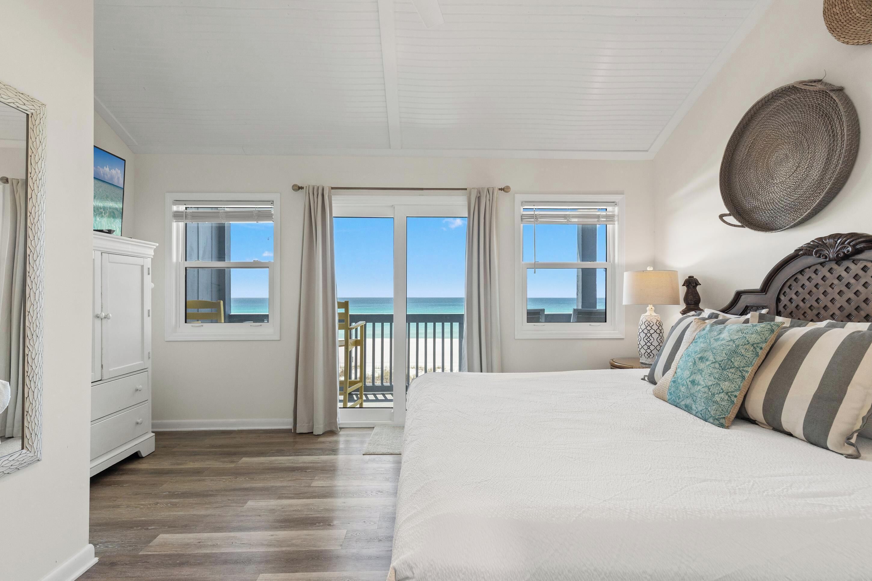 A bedroom with a large bed, a view of the ocean, a dresser, and a lamp.