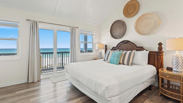 A bedroom with a large bed, a nightstand with a lamp, a wall clock, and a view of the ocean through a sliding door. - Premier Townhomes by Panhandle Getaways (Panama City Beach)
