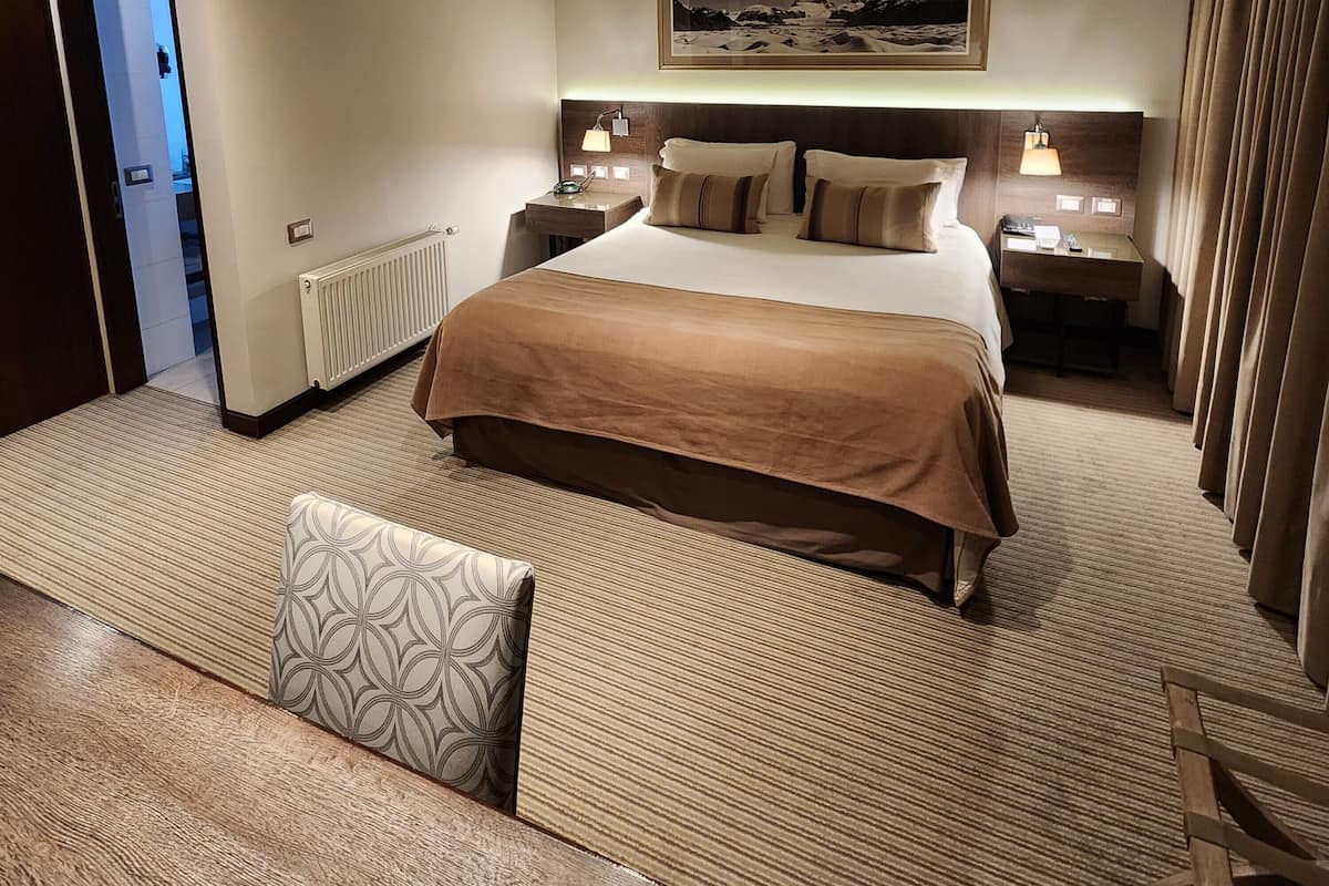 A hotel room with a large bed, two bedside tables, a headboard with a mounted artwork, a radiator, a patterned rug, and a bench with a pillow.
