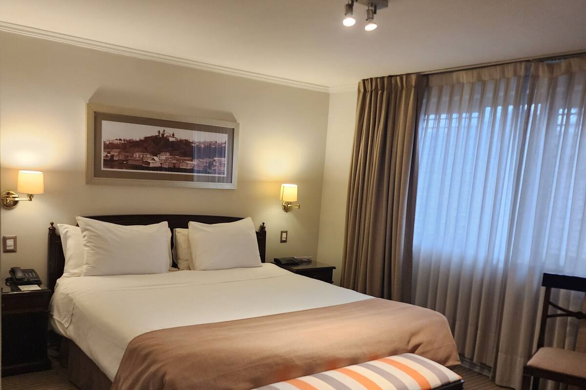 A hotel room with a large bed, two bedside lamps, a framed picture on the wall, a window with curtains, and a chair.