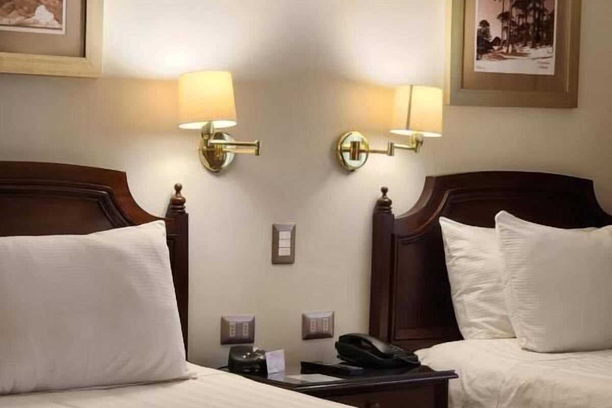 A hotel room with two beds, each with white linens and brown bedspreads. There are wall-mounted lamps on either side of the beds, and two framed pictures above the beds.