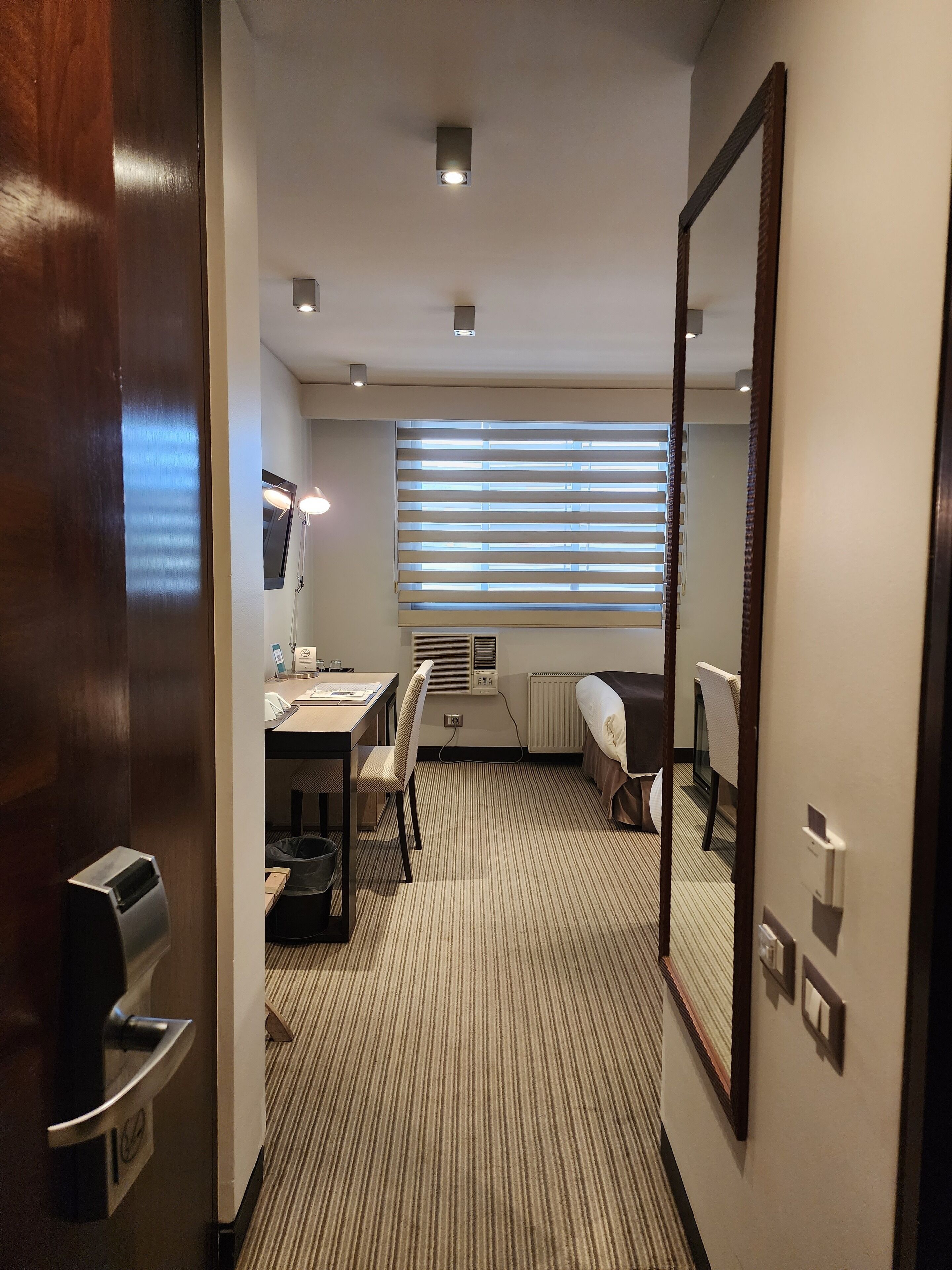 A hotel room with a large mirror, a desk with a chair, a radiator, and a window with blinds.
