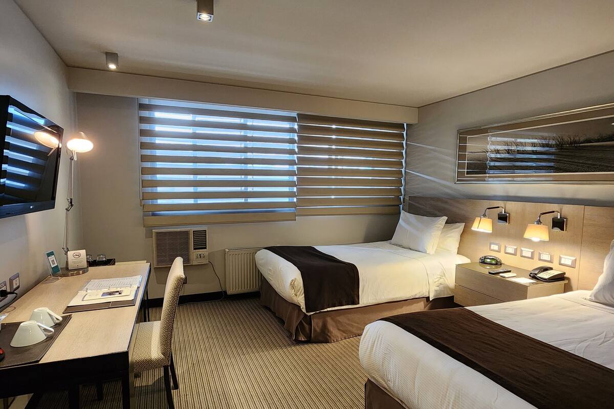 A hotel room with two beds, a desk, a chair, a TV, and a window with blinds.