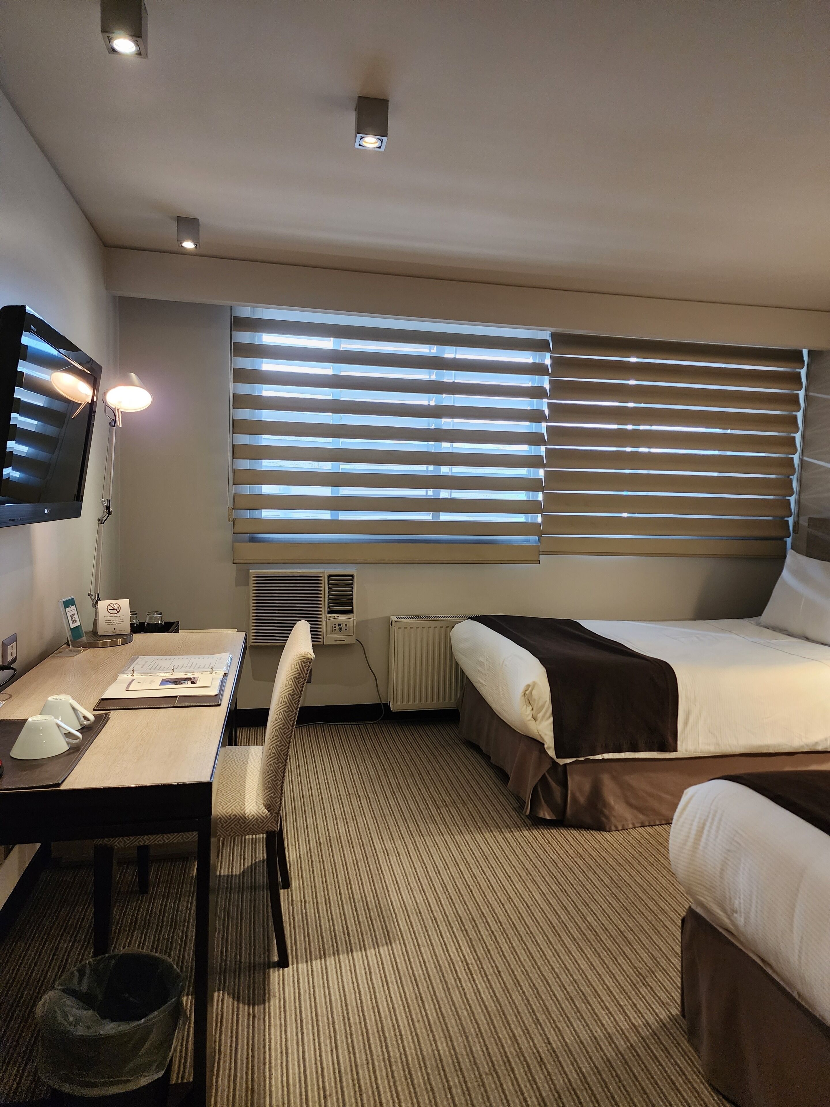 Quarto de hotel com cama, escrivaninha, cadeira, televisão e janela com persianas.