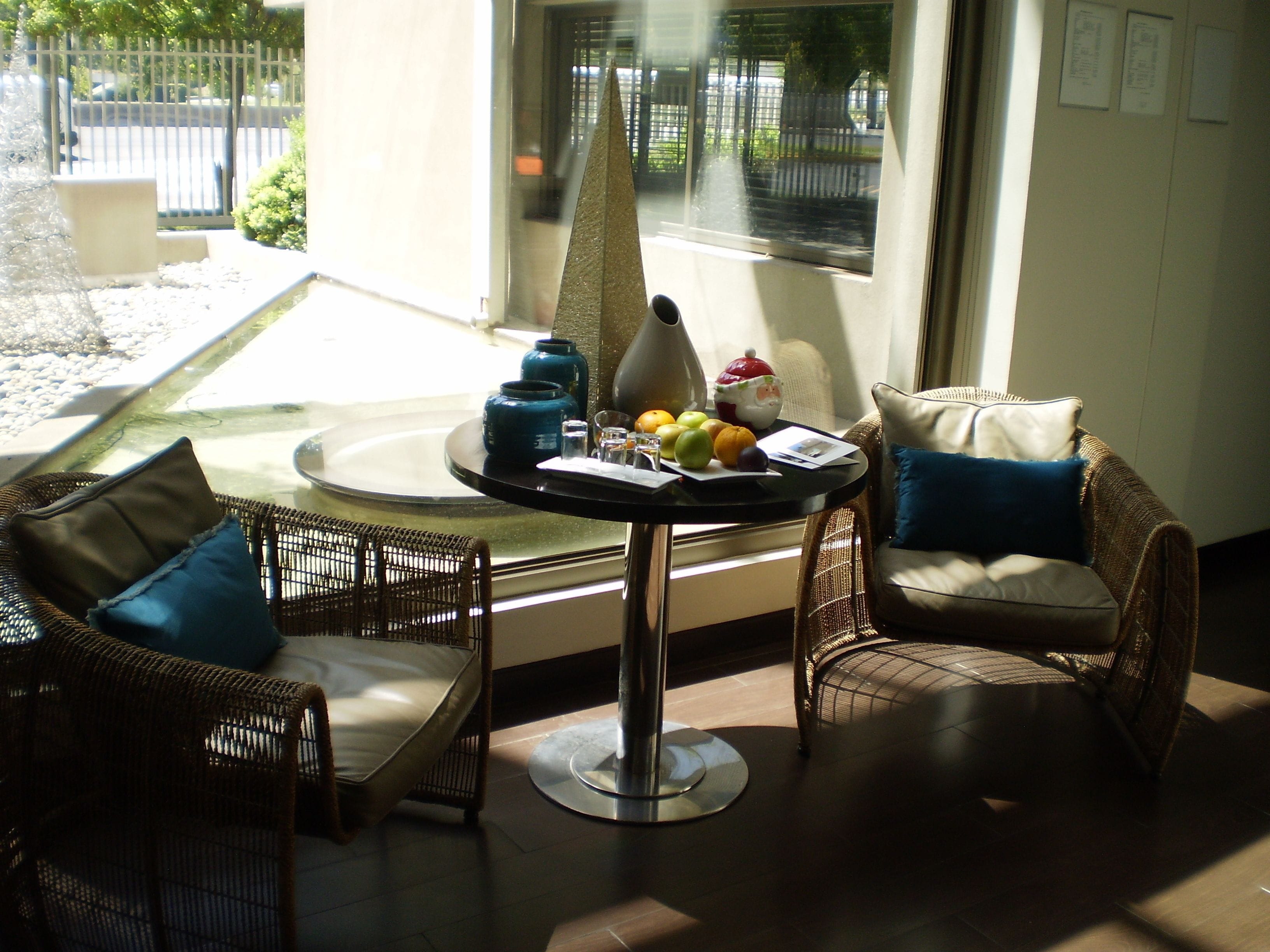 A sunlit room with wicker furniture, a round table with a vase, fruit, and papers, and a view of a street through a window.