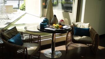 A sunlit room with wicker furniture, a round table with a vase, fruit, and papers, and a view of a street through a window.